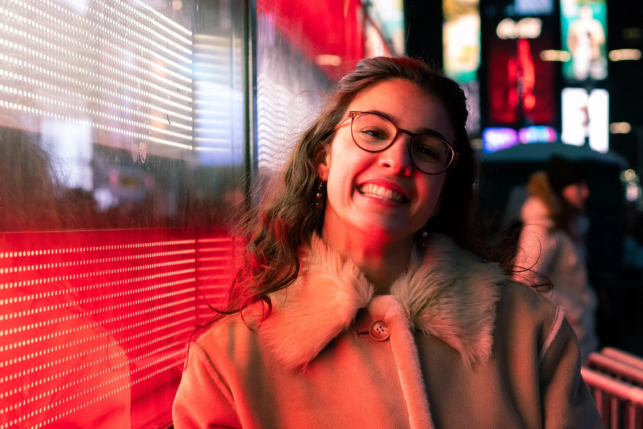 A smiling young woman with glasses and curly hair, wearing a fur-trimmed coat, stands next to a brightly lit red LED display in an urban setting at night.