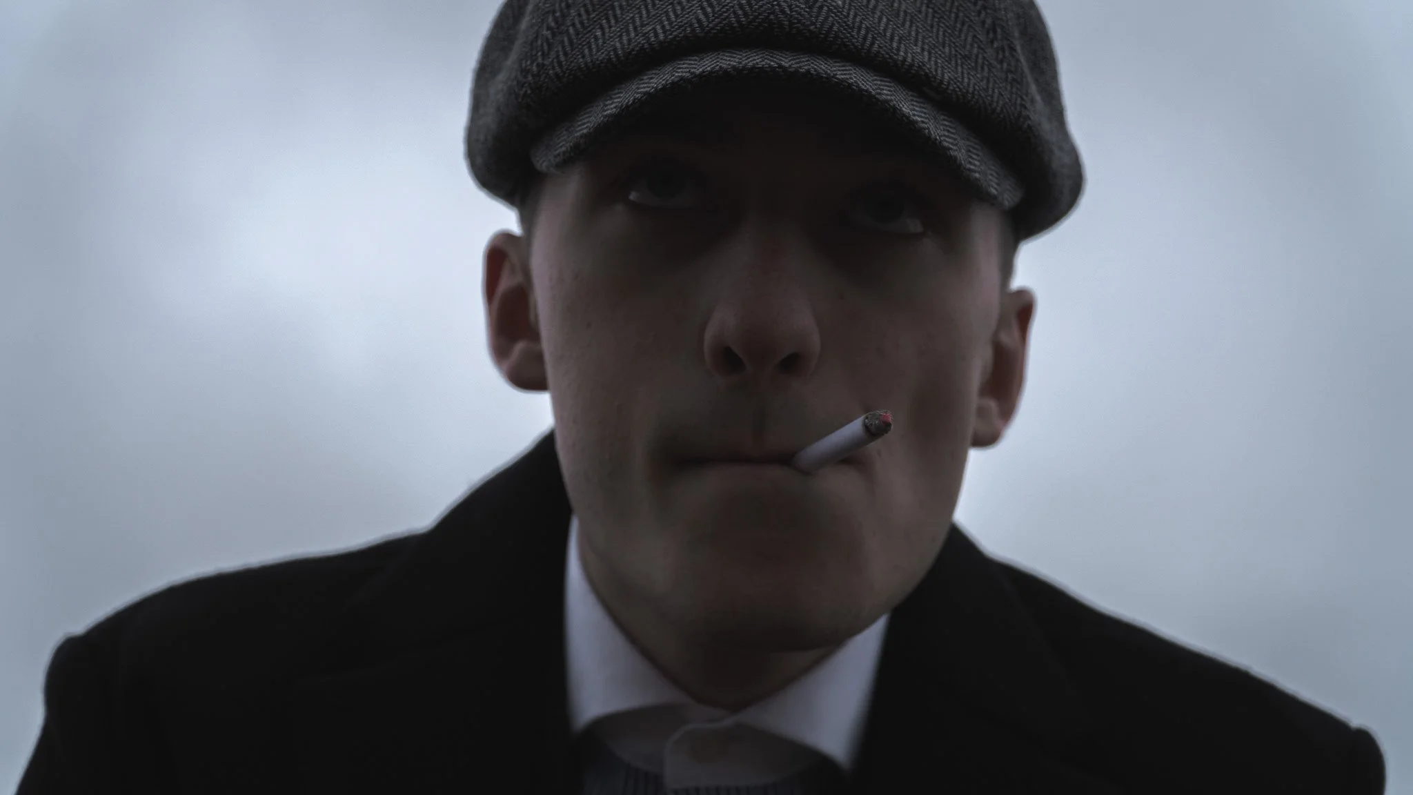 A young man with a serious expression, wearing a dark coat and a gray scarf, smokes a cigarette against a gray sky background.