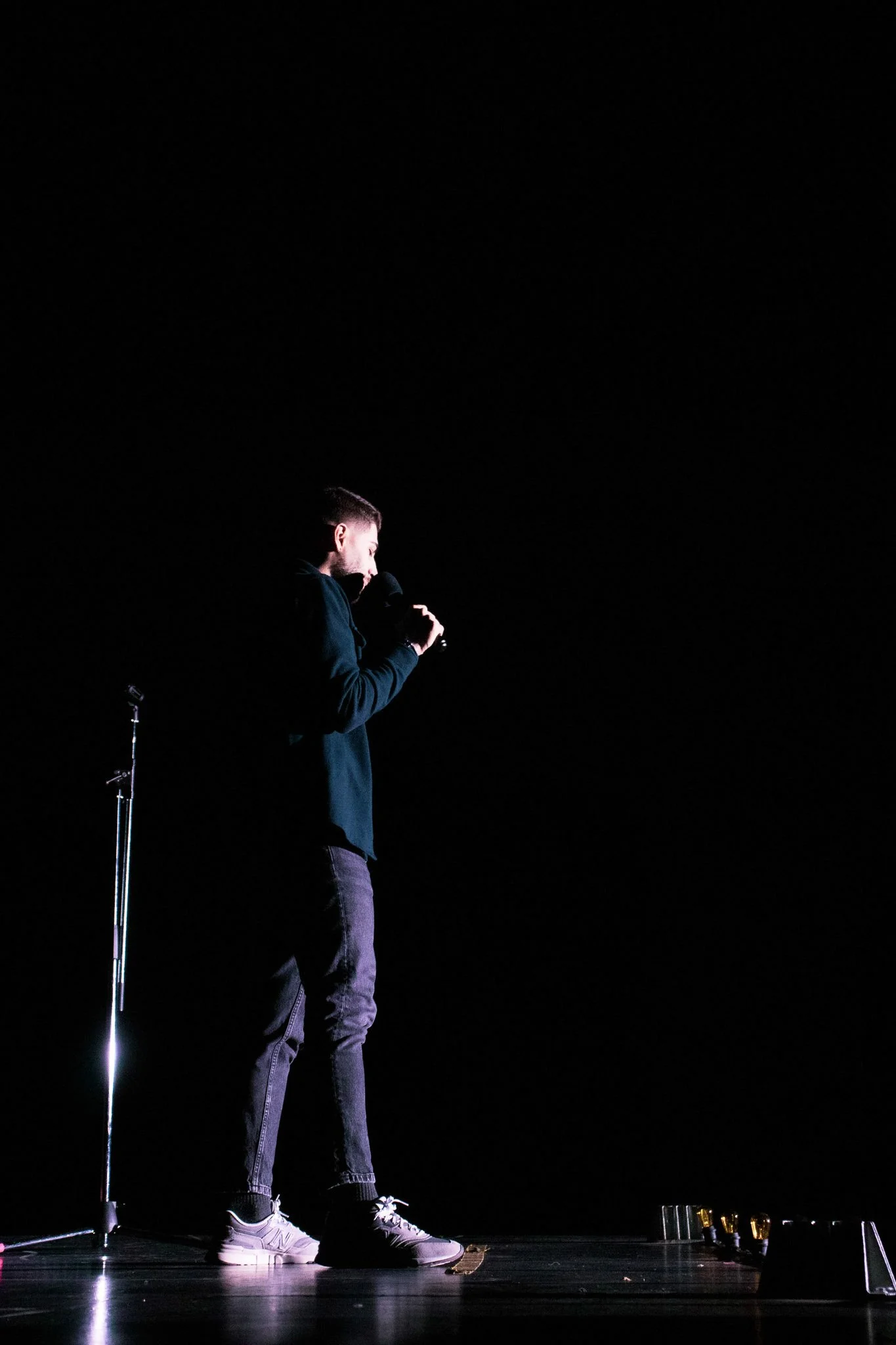 A man standing on a stage at night holds a microphone close to his mouth, dressed in a dark hoodie and pants, with white sneakers, illuminated by a spotlight against a dark background.