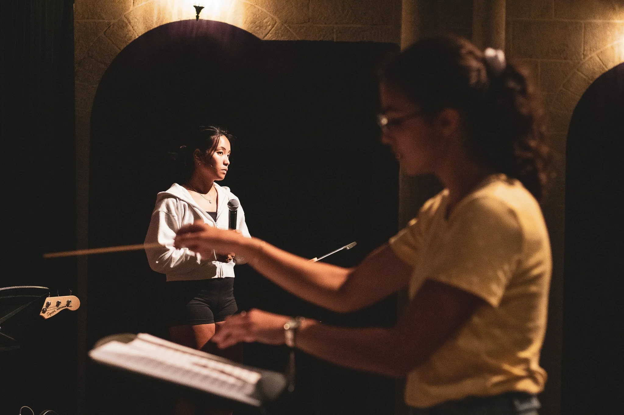 A young woman stands with a microphone in her hand during a performance or rehearsal, with another person playing the piano in the foreground. The scene is dimly lit, with a dark background and warm lighting.