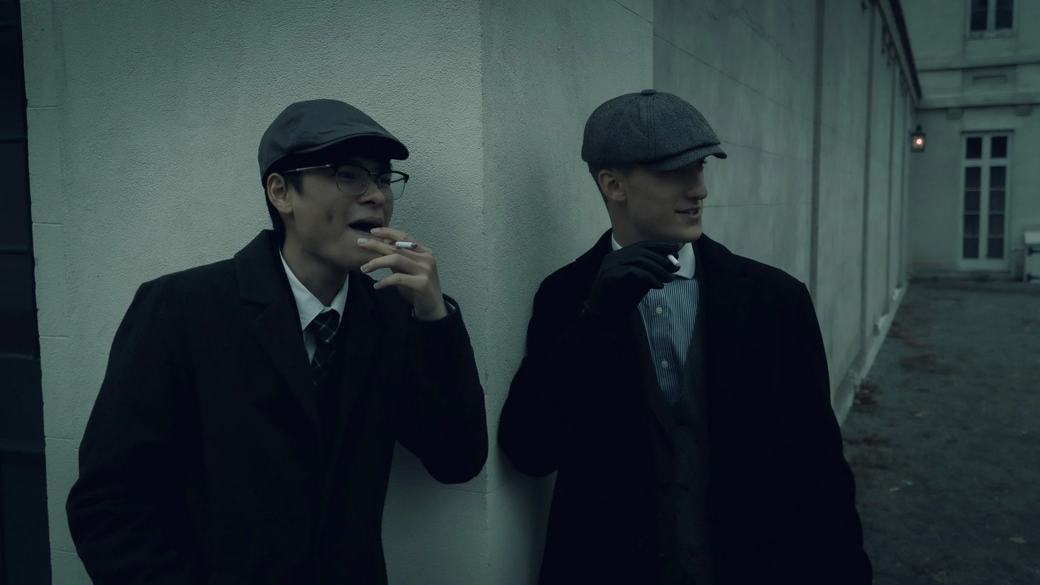 Two men in vintage suits and hats standing against a white wall on a cold, overcast day. One is smoking a cigarette, and the other is holding a cigarette to his mouth.