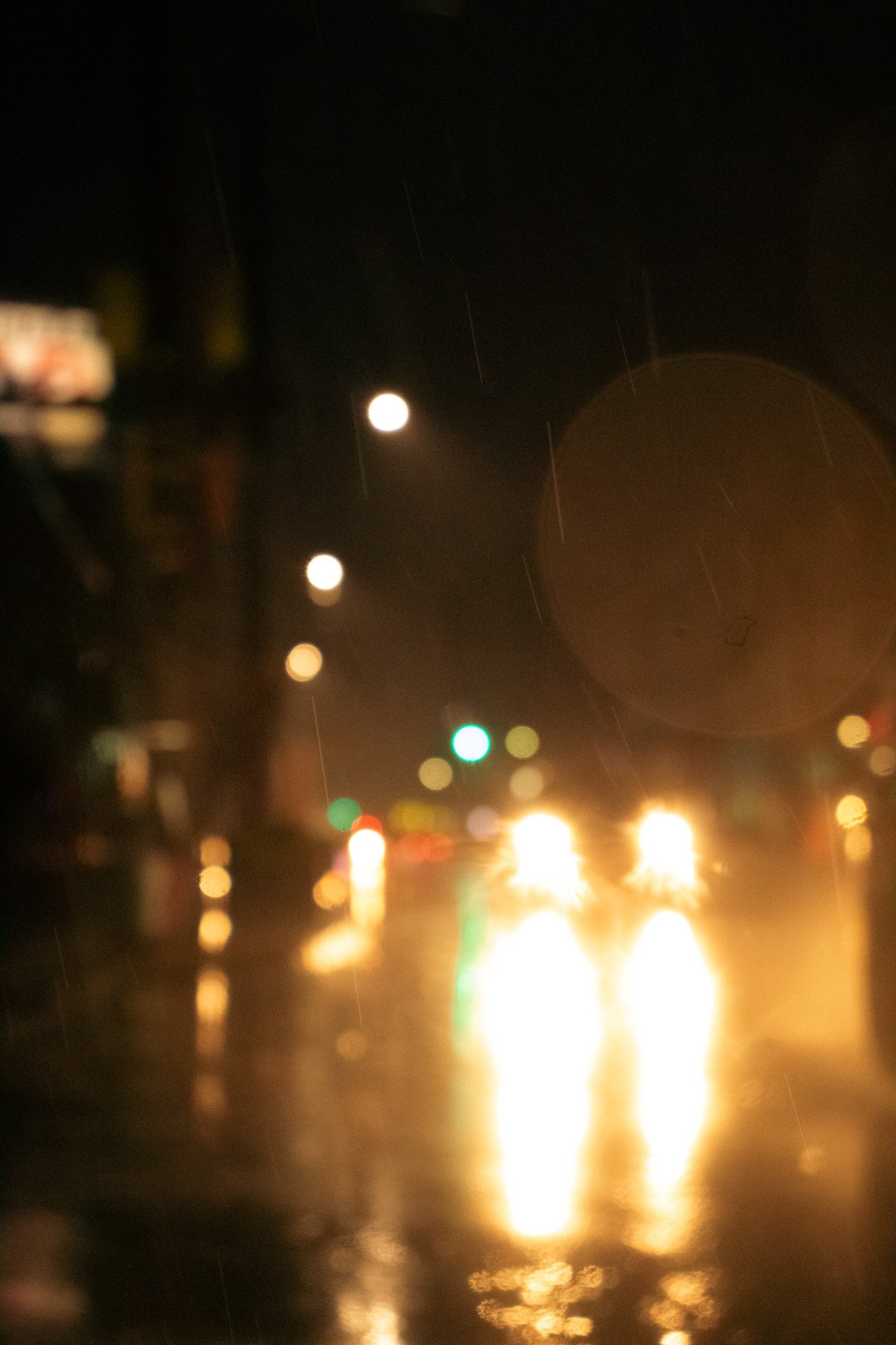 A blurry, nighttime scene of a street with colored lights reflecting on wet pavement, with rain falling.
