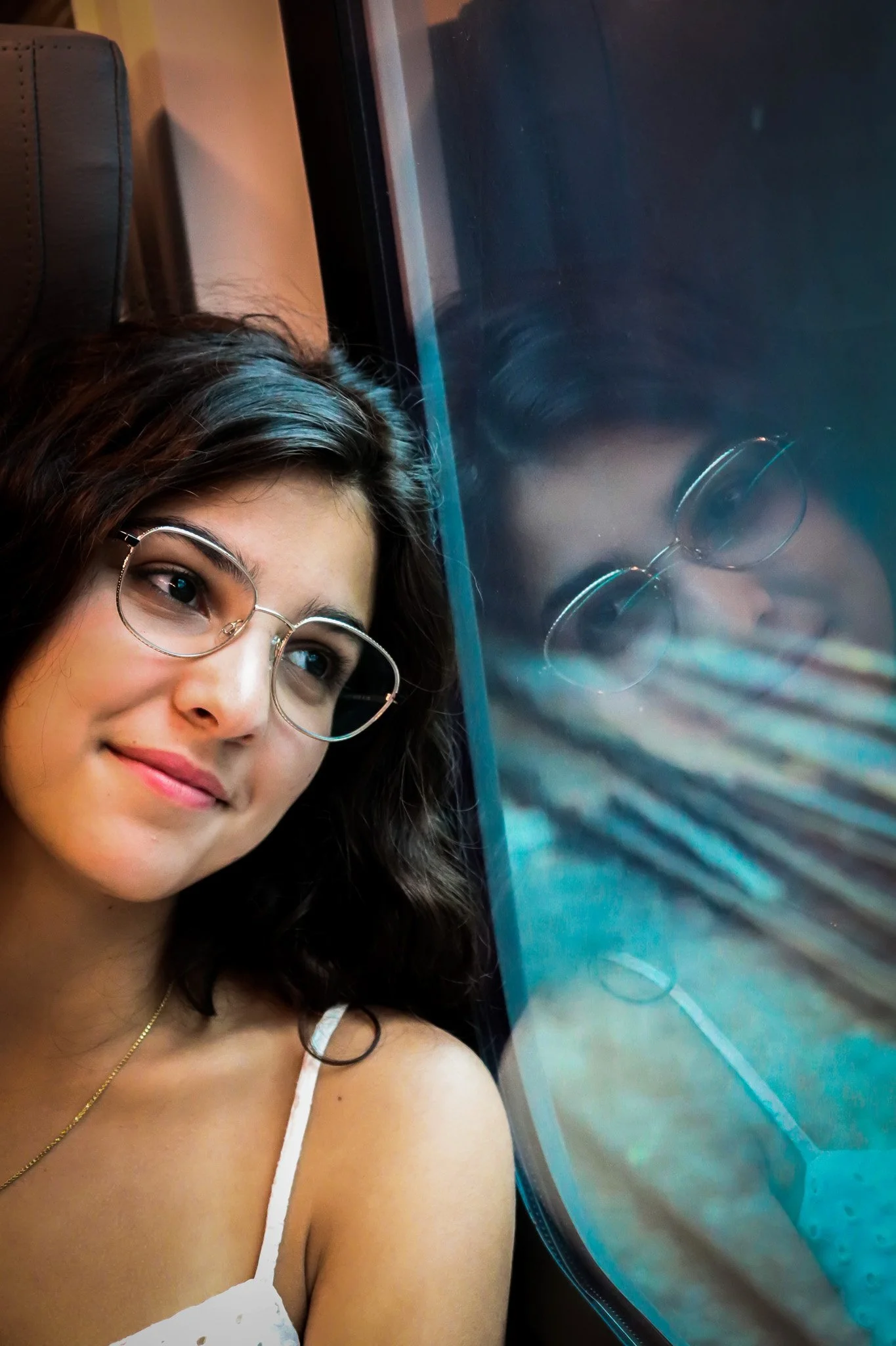 A young woman with glasses and dark hair is smiling while taking a selfie, with a large screen displaying an abstract image of water and ripples behind her.