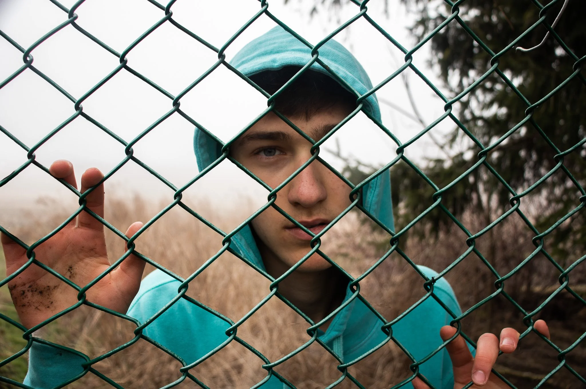 A young man in a blue hoodie looking through a chain-link fence.
