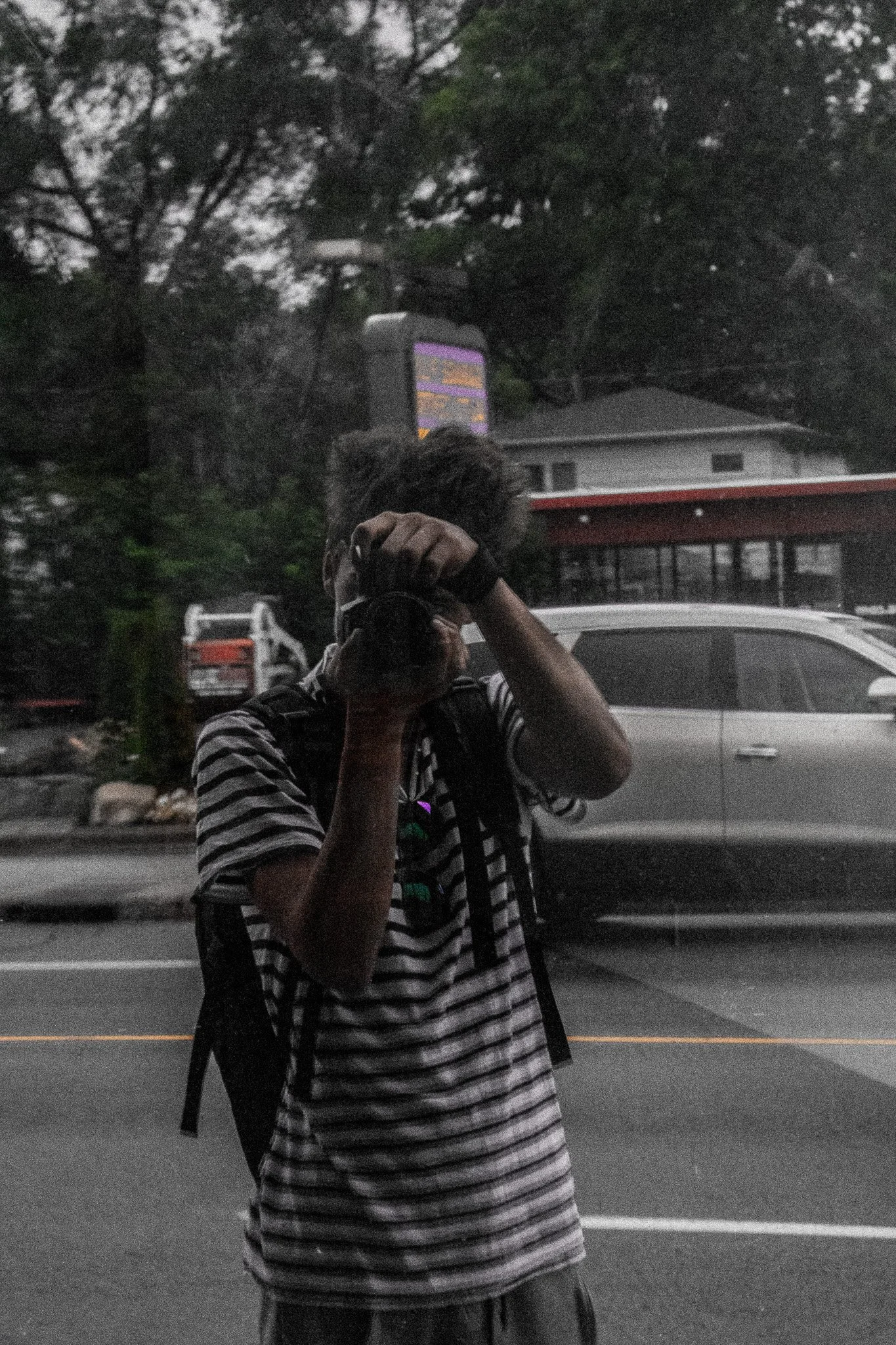 A person in a striped shirt is taking a photo of their reflection in a mirror while in a parking lot. The person's face is not visible, and they are wearing a backpack. The background includes trees, a house, and parked cars, with the scene appearing