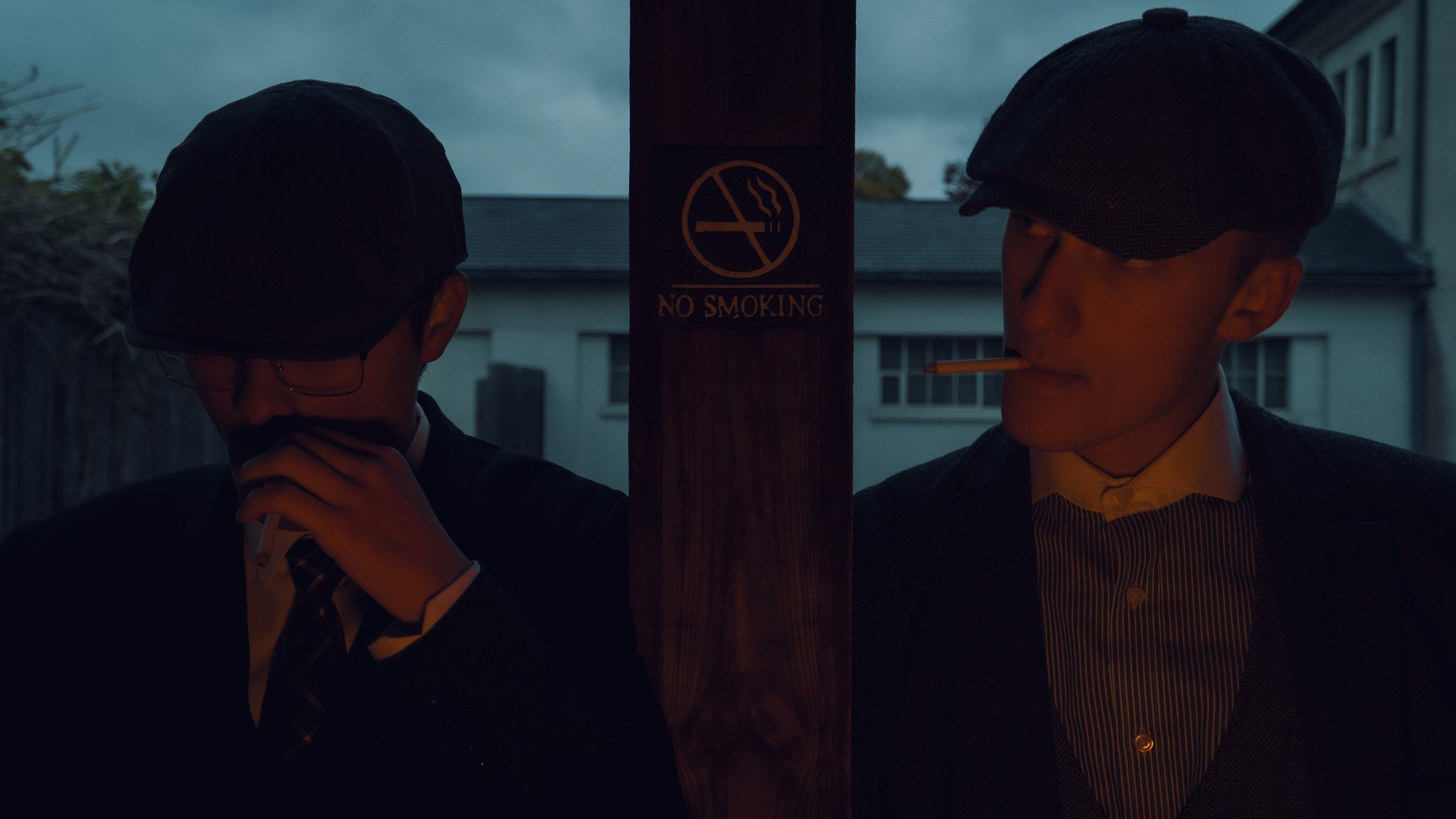Two people stand on opposite sides of a wooden pole with a "No Smoking" sign, one person on the left adjusting their glasses and the other person on the right holding a cigarette in their mouth, both wearing dark clothing and hats outdoors during the