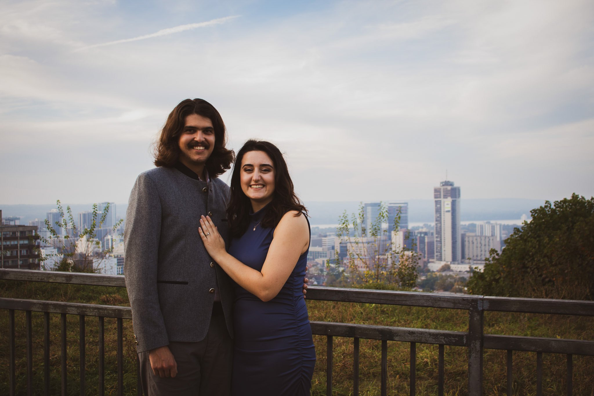 A smiling young couple stands closely together outdoors with a city skyline in the background, under a partly cloudy sky.