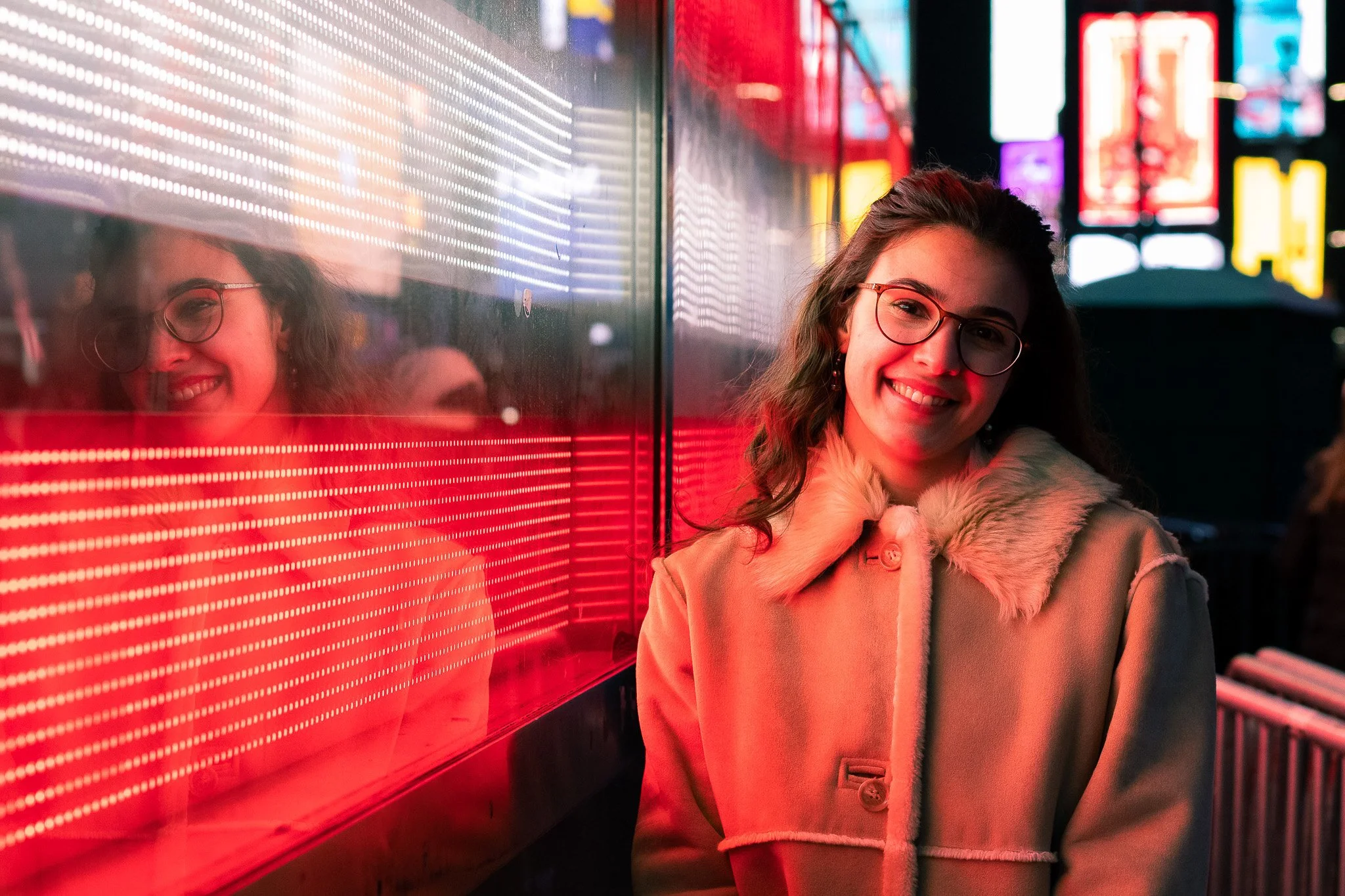 A young woman with glasses and long, wavy hair, smiling in a neon-lit urban setting at night, standing beside a brightly lit digital display with colorful neon signs in the background.