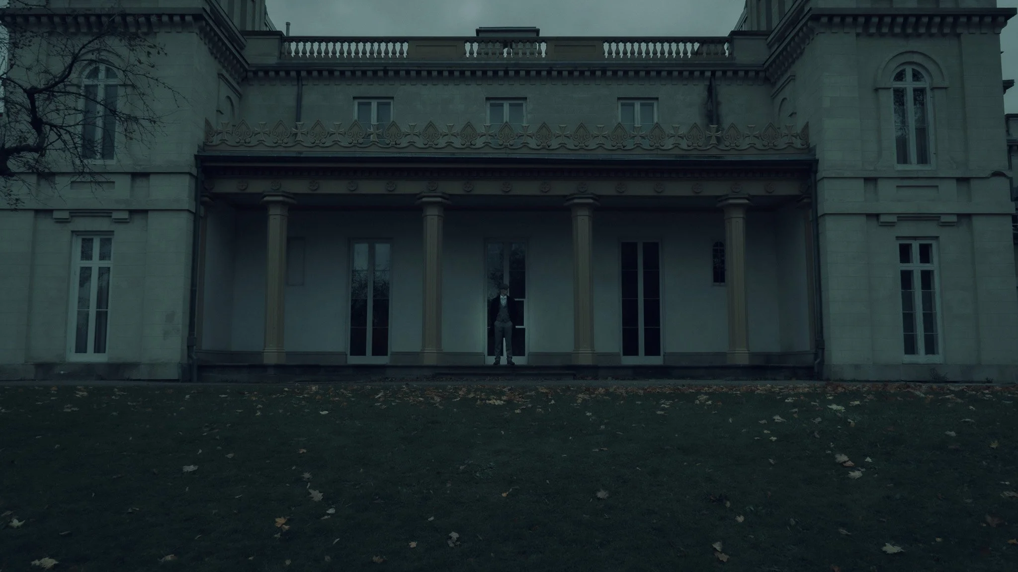 A man dressed in formal attire stands on the porch of a large historic mansion with ornate architectural details, columns, and multiple windows. The scene appears to be overcast and there are fallen leaves on the ground.