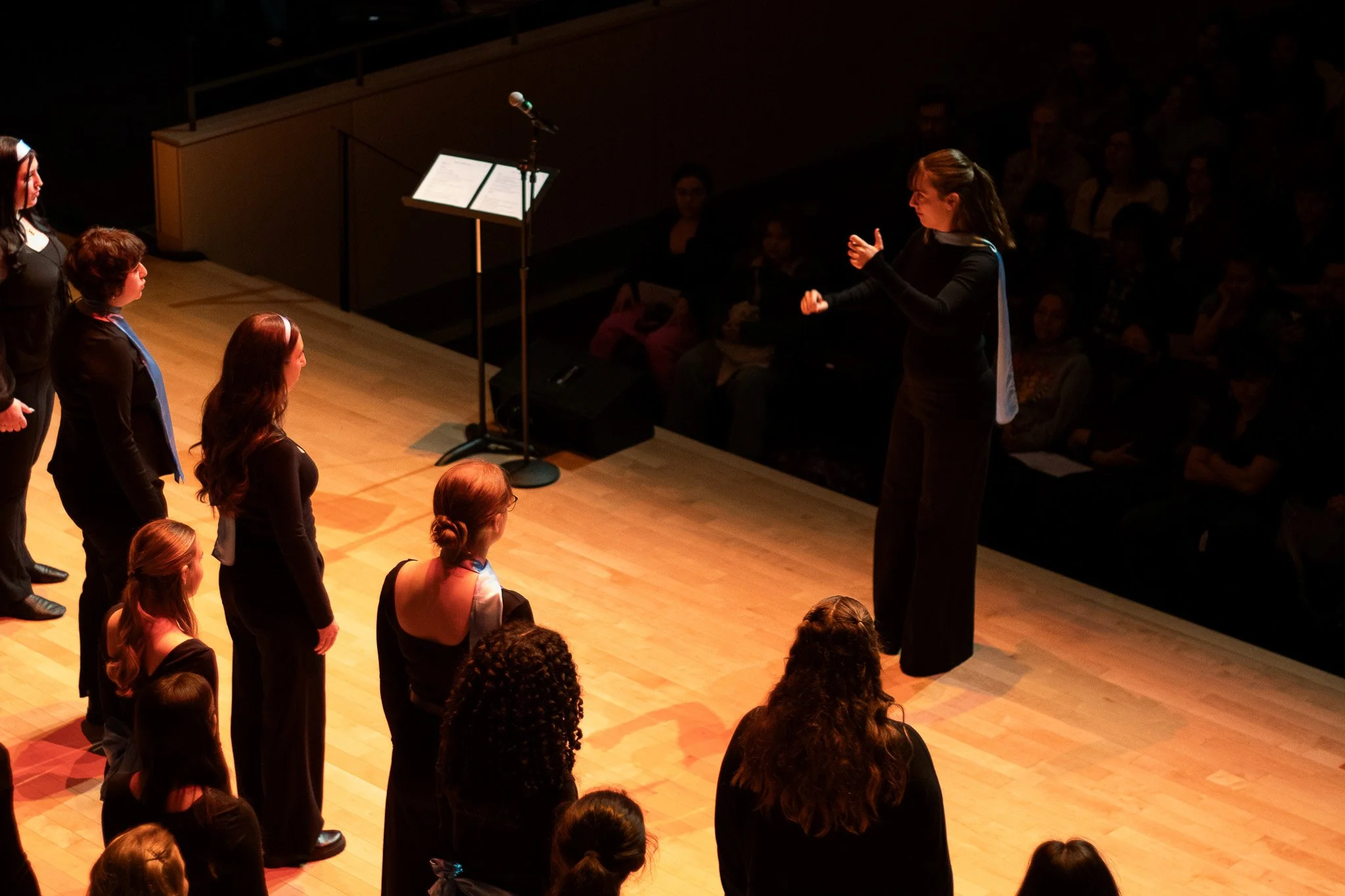 A woman standing on a stage appears to be conducting or directing a group of women facing her, with an audience watching in a dimly lit auditorium.