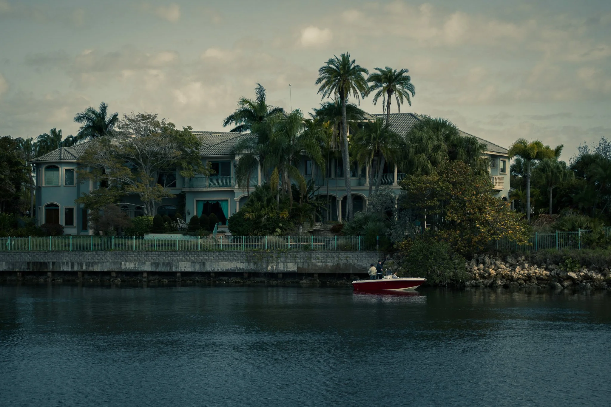 Luxury house with palm trees on waterfront, with a boat in the water, overcast sky.