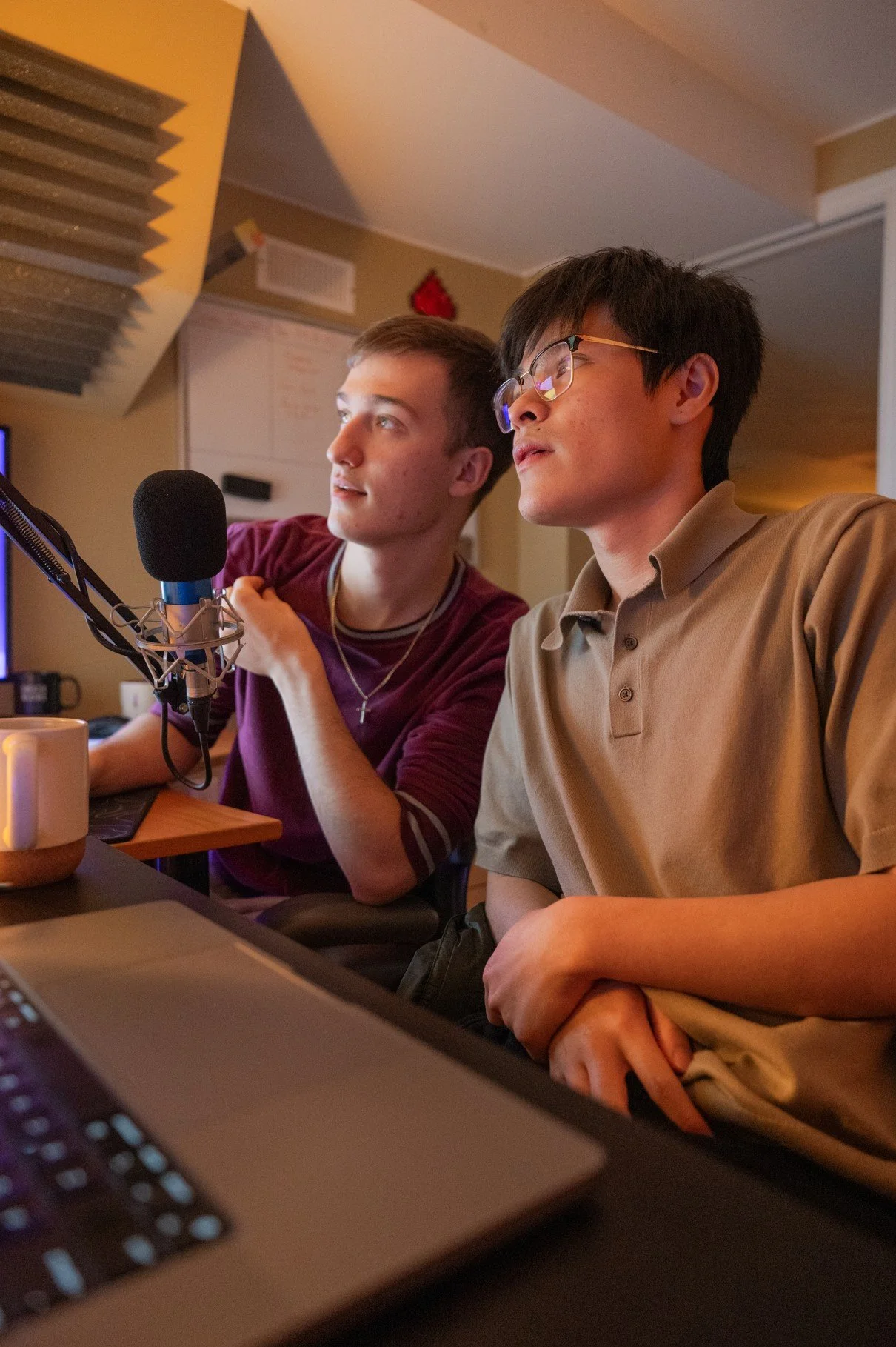 Two young men sitting at a desk with a microphone, monitoring a computer screen, in a room with soundproofing and a whiteboard.