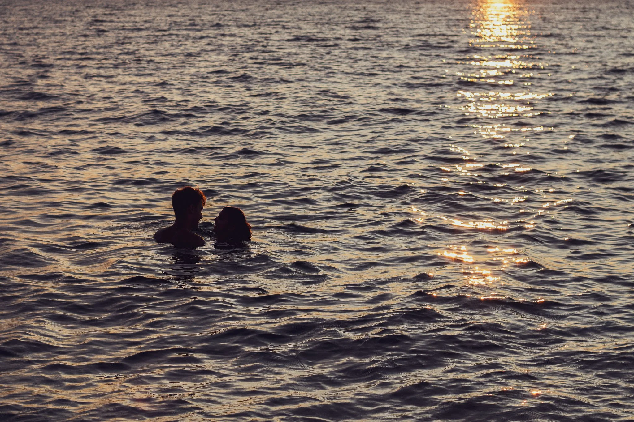 Two people are swimming in the ocean during sunset, with sunlight reflecting off the water.