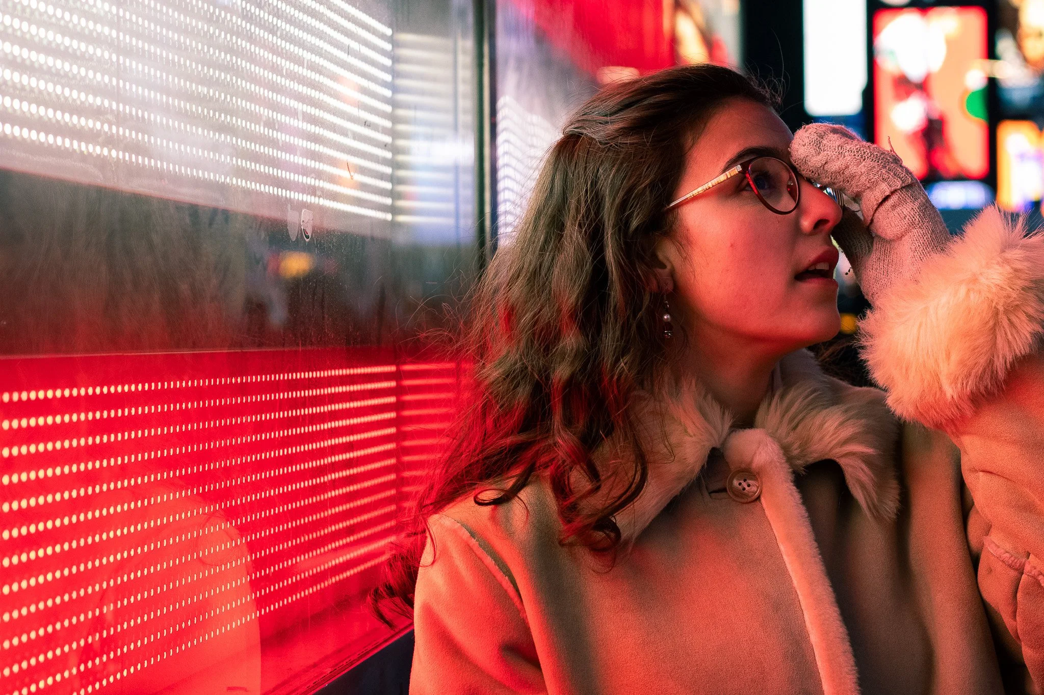 A young woman with glasses and earrings, wearing a furry jacket, touching her forehead in front of a red illuminated display on a wall, with colorful bright screens in the background.