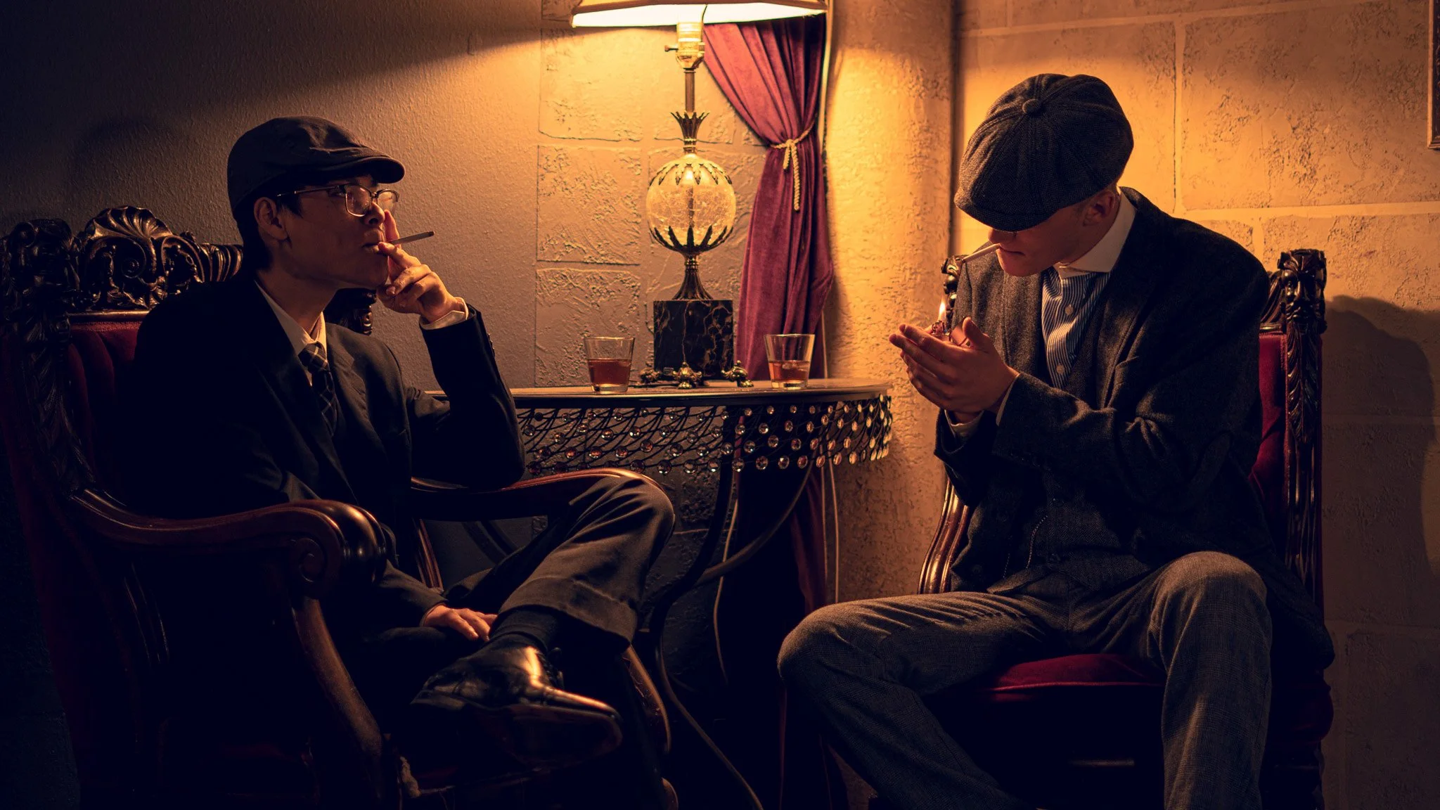 Two men in vintage-style clothing sitting in a dimly lit room with ornate chairs and a table, one smoking a cigarette and the other looking at a phone.