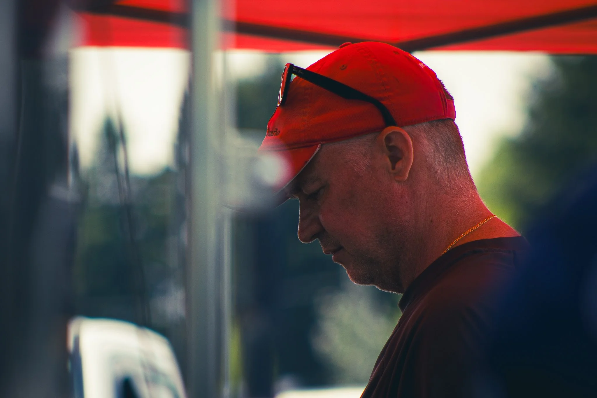 A person wearing a red cap and sunglasses resting on the cap, looking downward with a neutral expression.