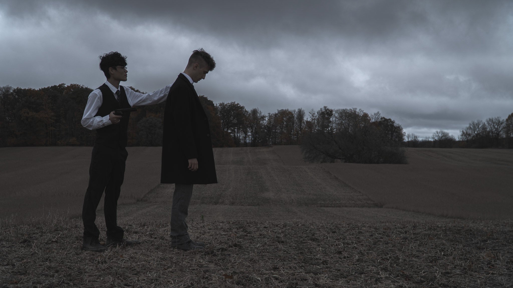Two young men in suits standing on a dirt path in a rural area on a cloudy day. One man touches the other's shoulder and is holding a phone. The background features trees and a gloomy sky.