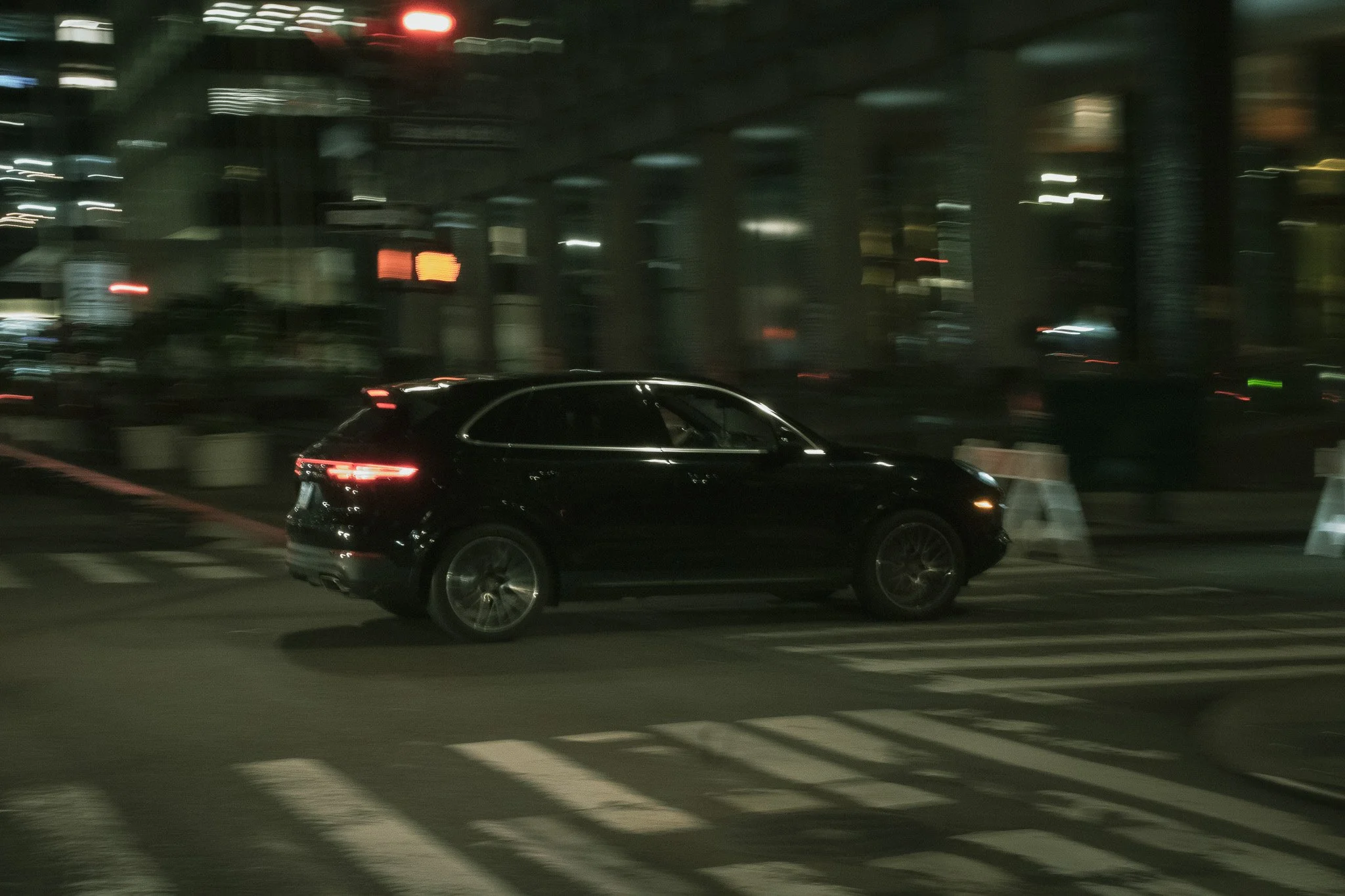 A black car driving past a city street at night with blurred lights and buildings in the background.