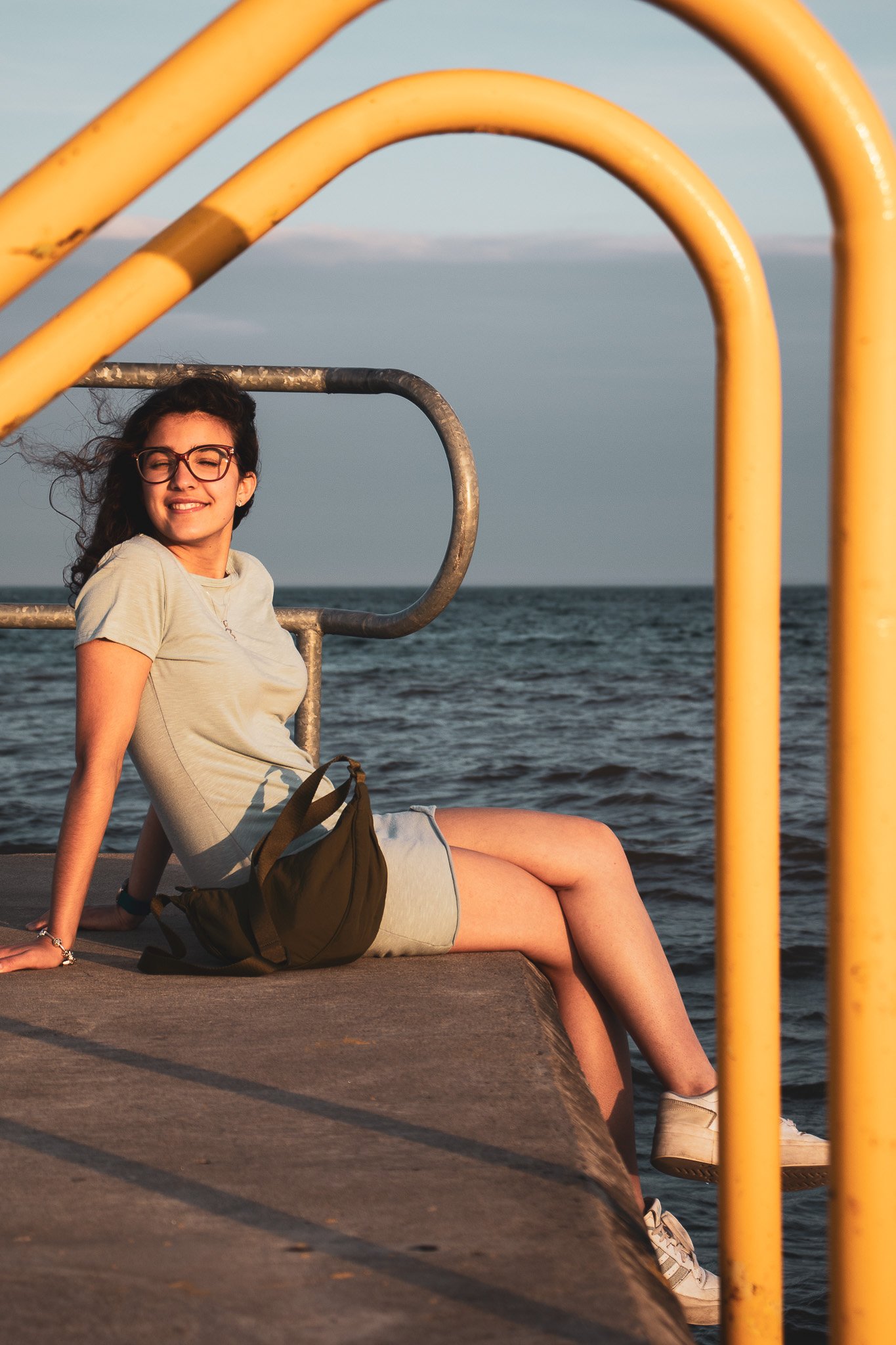 A young woman with dark curly hair, wearing glasses and a light beige t-shirt, sitting on a dock by the water during sunset or late afternoon. She is smiling with her eyes closed, and her legs extend over the edge of the dock. Yellow railing bars are