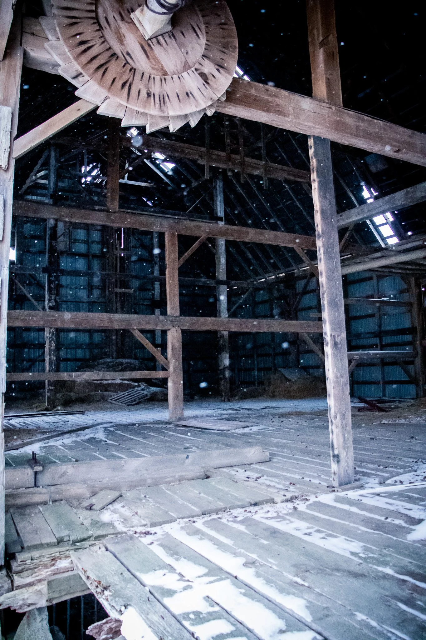 Interior of an unfinished wooden attic with exposed beams and snow-covered floor, showing sunlight through gaps in the roof.