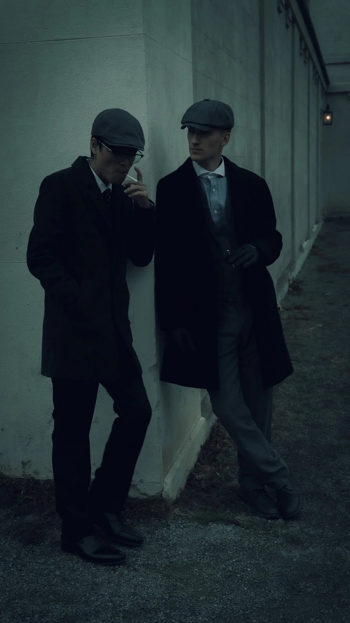 Two men wearing dark suits, hats, and gloves standing against a concrete wall with a dimly lit background, one smoking a cigarette.