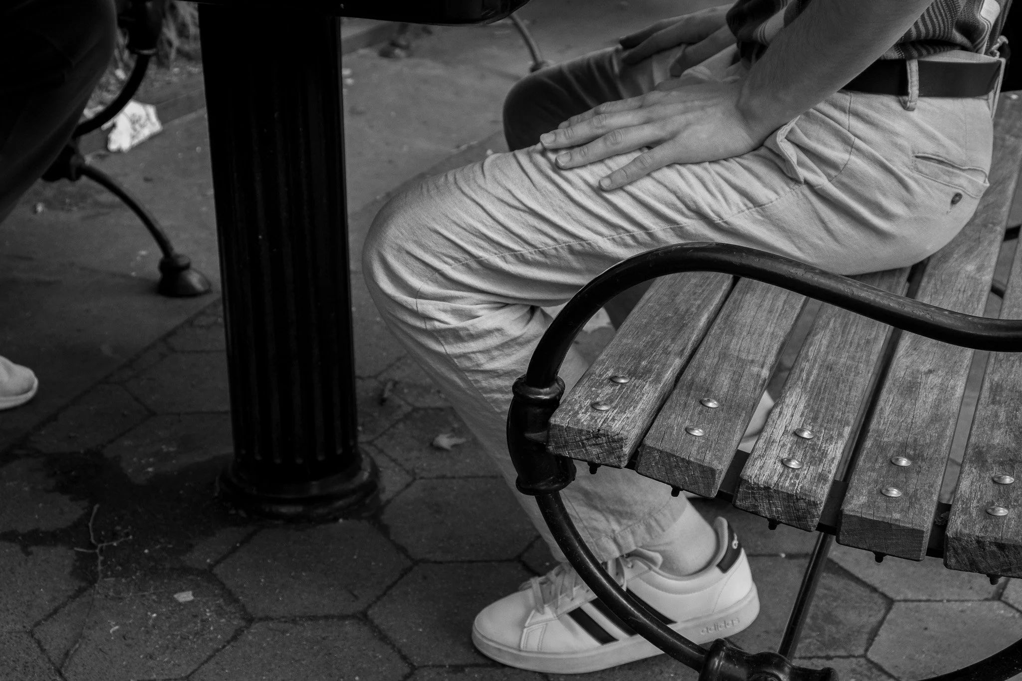 A person wearing light-colored pants and white sneakers sitting on a wooden park bench with their hand resting on their thigh. The image is in black and white and part of another person is visible on the left side.