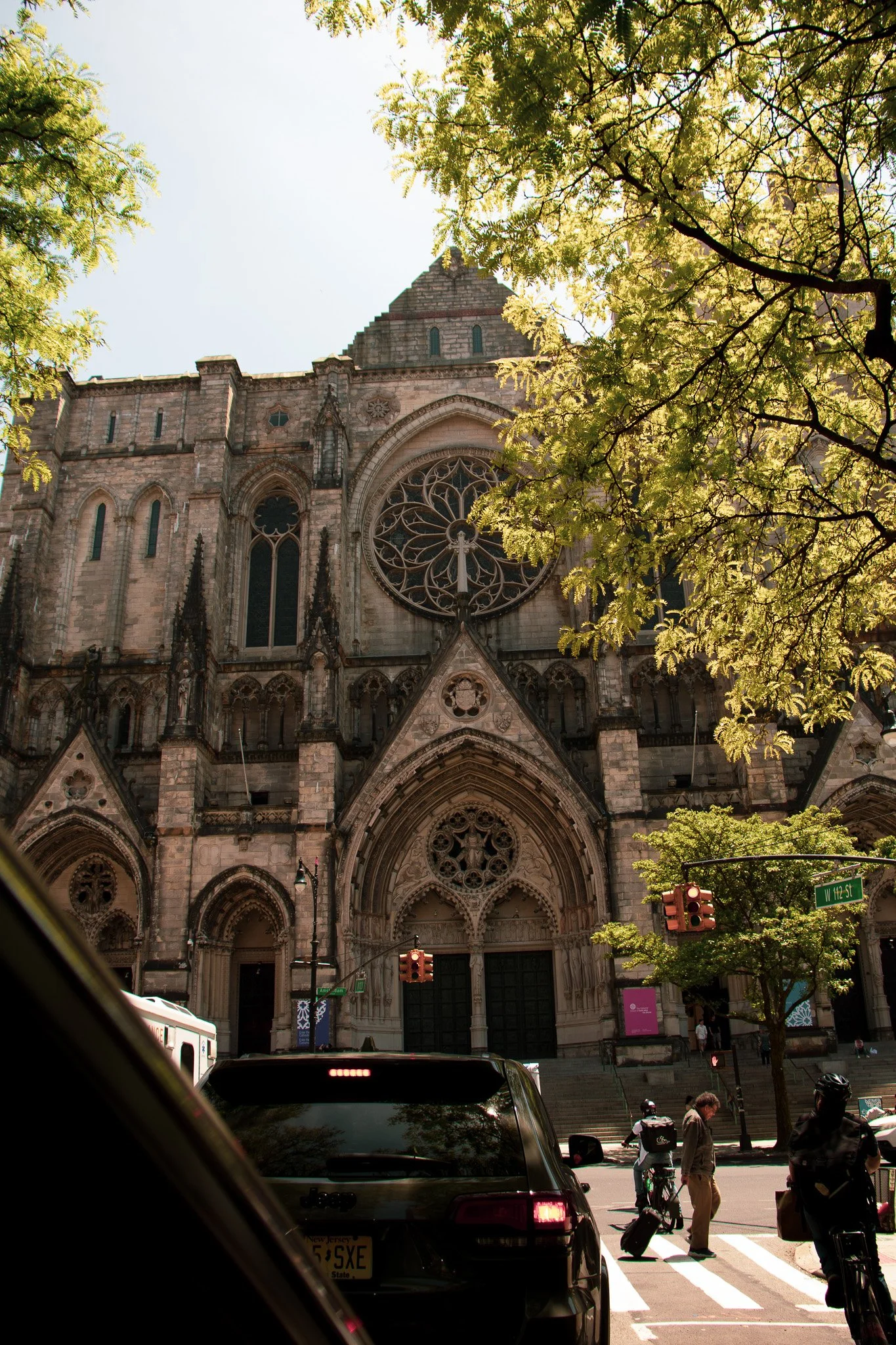 The front facade of a historic stone Gothic-style church with stained glass windows and a large rose window, surrounded by trees, with people walking and cars stopped at a street corner.