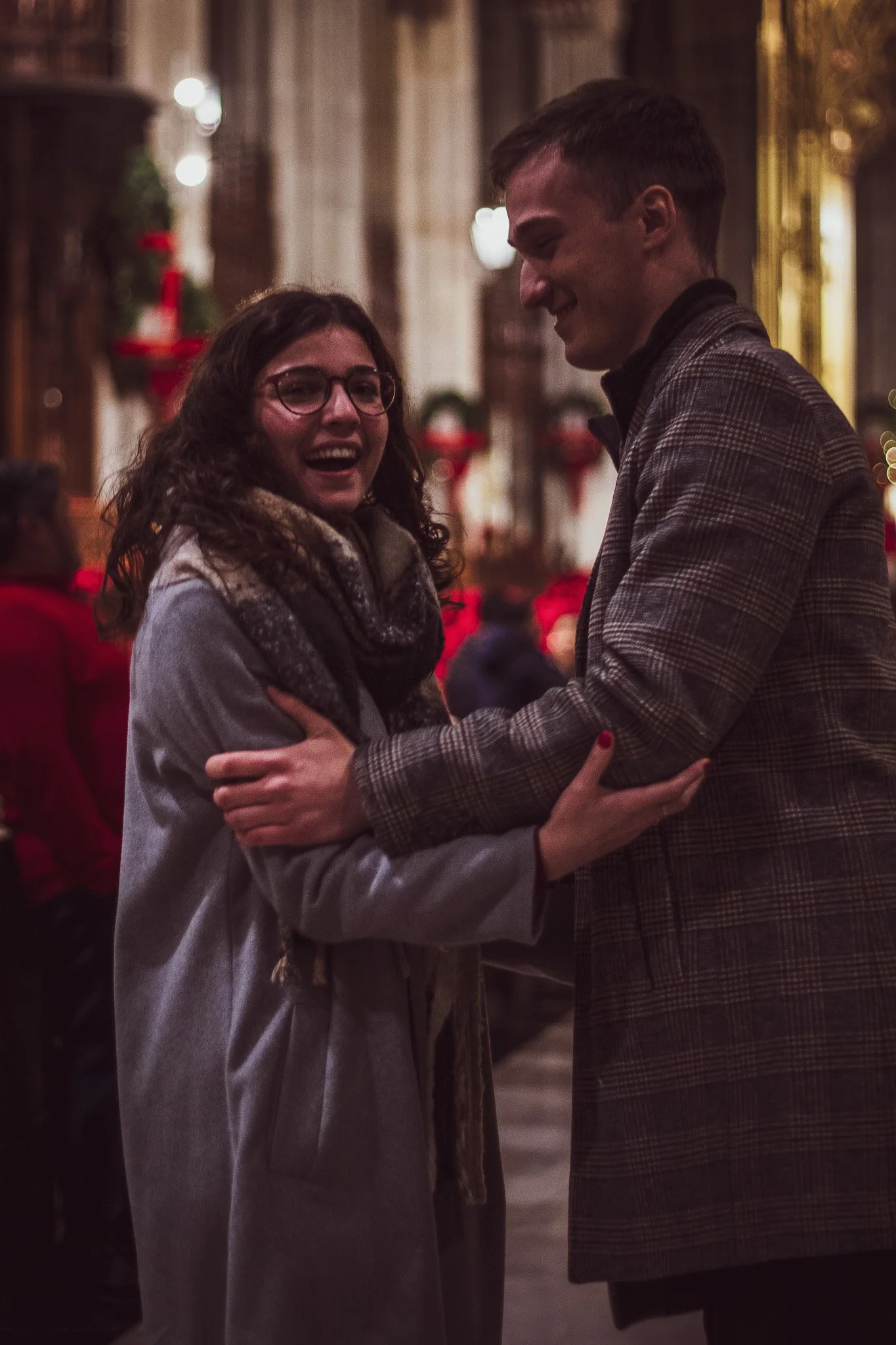 A young woman with curly hair and glasses smiling, holding hands with a young man in a plaid jacket, both sharing a joyful moment indoors with warm lighting and holiday decorations in the background.