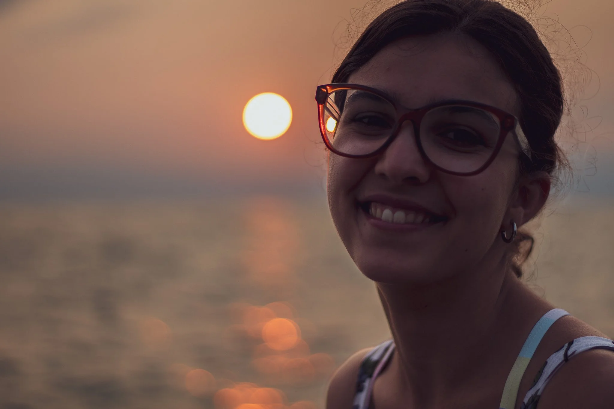 A woman smiling outdoors at sunset, wearing glasses and a sleeveless top, with the sun partially visible behind her.