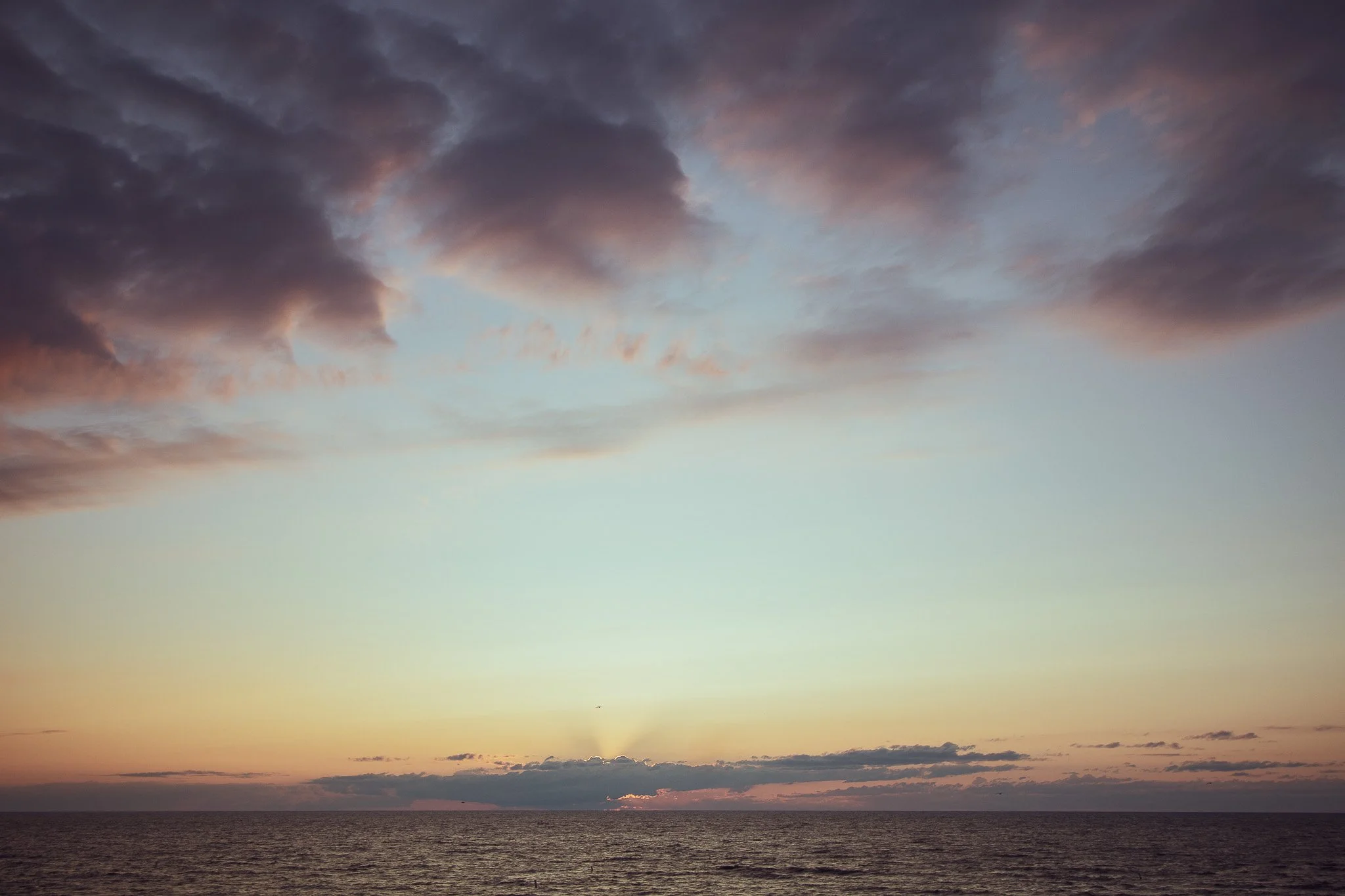 Sunset over the ocean with dark clouds and a faint glow of sunlight near the horizon.
