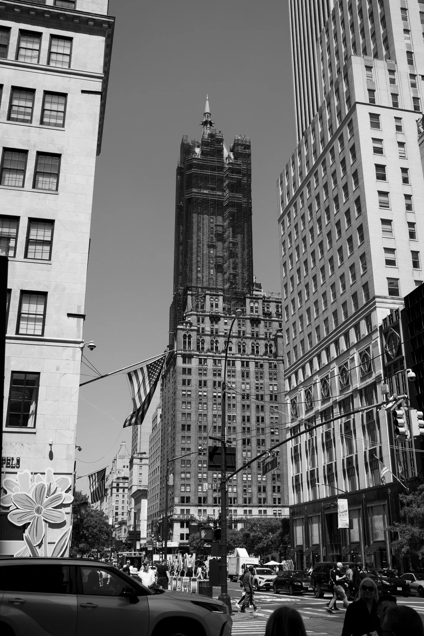 Black and white photo of busy city street scene with tall buildings, pedestrians crossing a crosswalk, and cars driving by. A construction site with scaffolding is visible up the central building.
