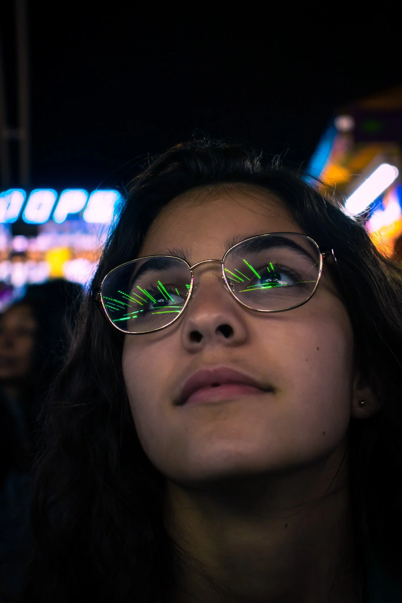 Close-up of a person's face with glasses, nighttime background with bright neon signs and blurry lights nearby.