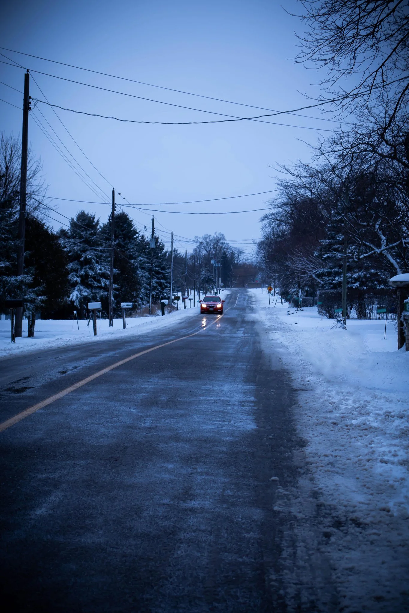 A snowy street scene at dusk with a single car driving towards the camera. Snow covers the ground and trees, power lines stretch across the sky, and a few mailboxes are lining the side of the road.