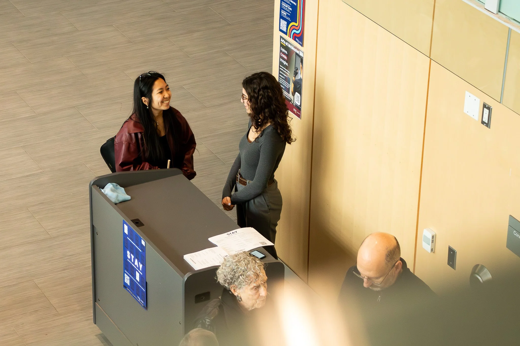 Two women are shaking hands and smiling at each other at a reception desk. One woman has long dark hair and wears a maroon jacket, while the other has curly brown hair and glasses and wears a gray sweater. There are papers, a mobile phone, and a sign