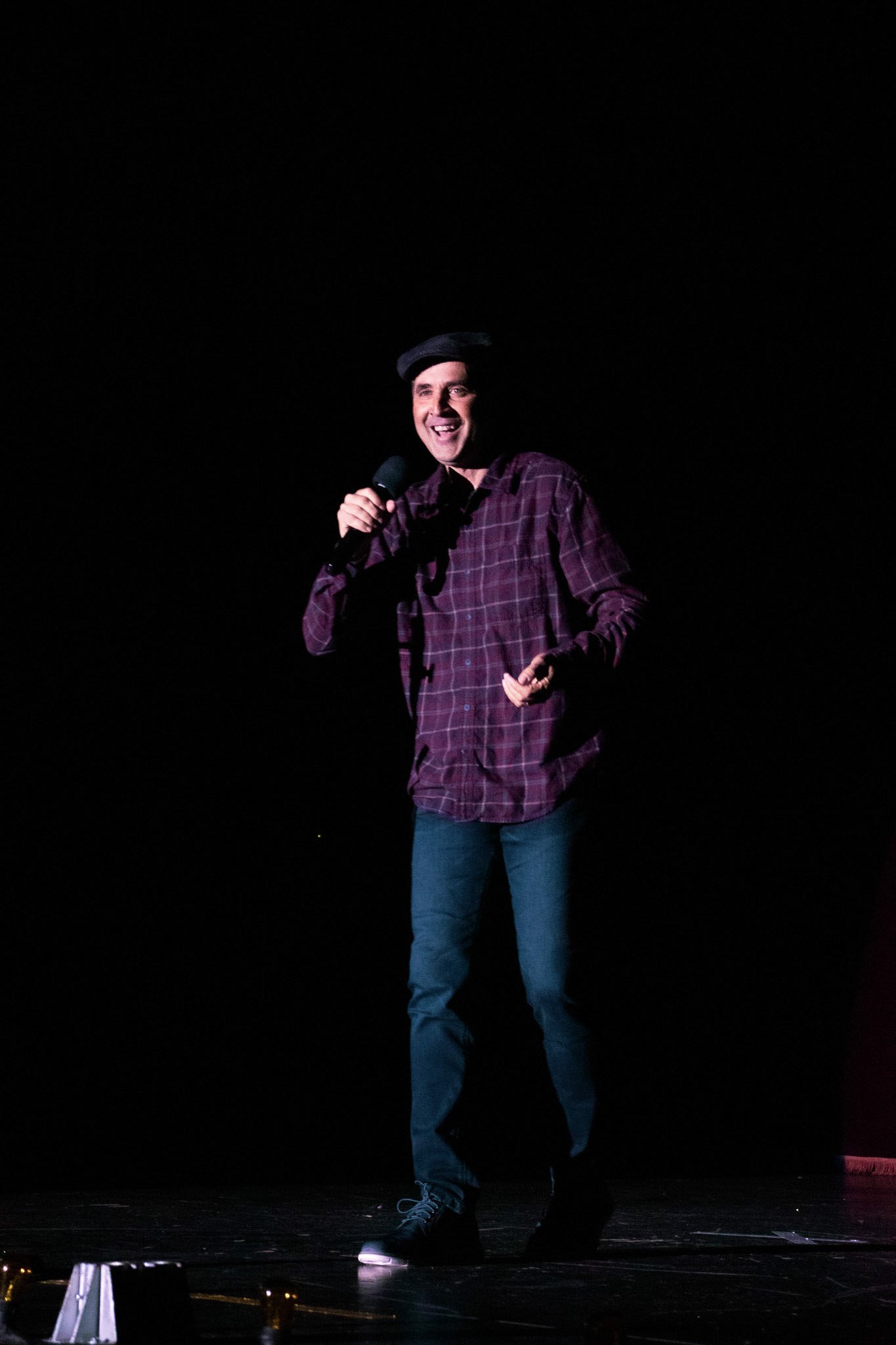 Man performing stand-up comedy on stage, holding a microphone, wearing a black beret, plaid shirt, jeans, and sneakers, smiling and laughing.