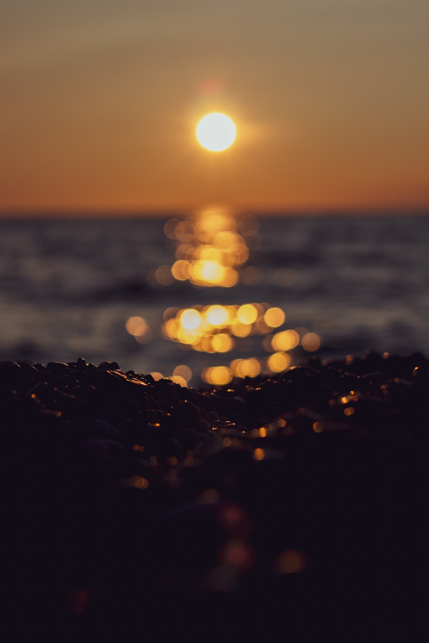 A sunset over the ocean with the sun low on the horizon and its reflection on the water, viewed from a rocky shoreline.