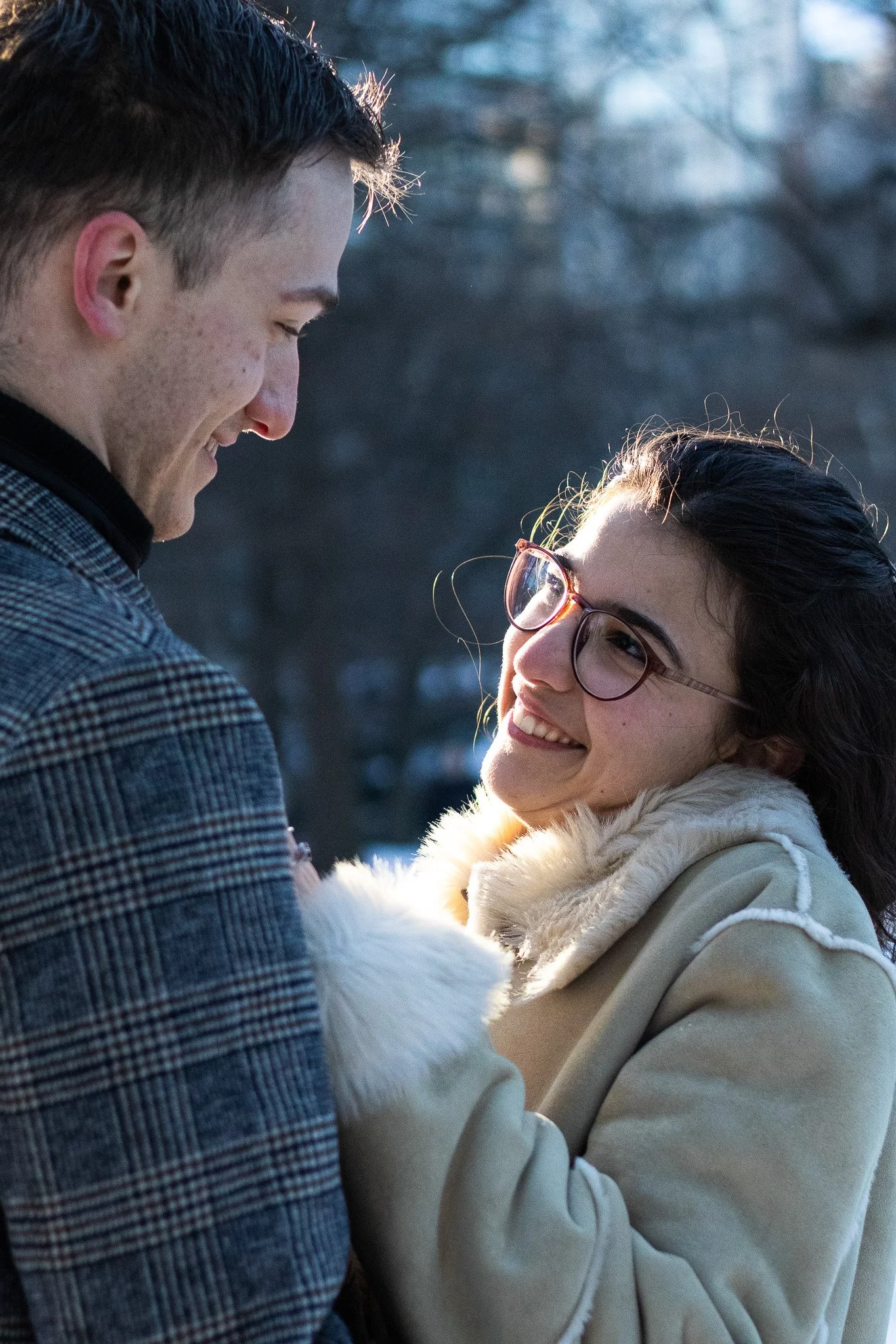A smiling couple is outdoors, sharing an intimate moment during winter, with the woman wearing glasses and a beige coat with a fur collar, and the man wearing a gray checkered coat.