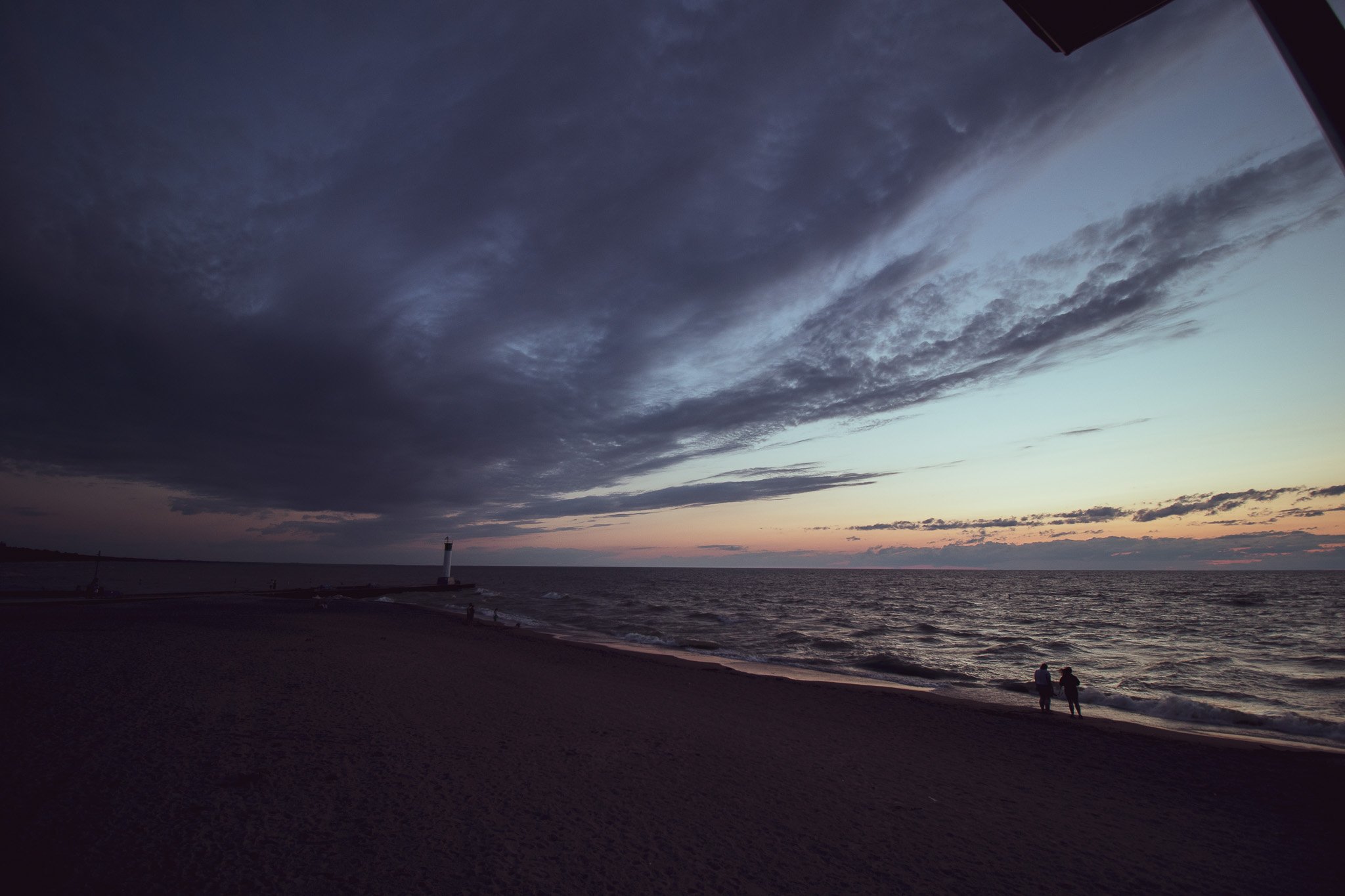 A sunset or twilight over a beach with dark clouds in the sky. Two people are walking along the shoreline near the water, and a lighthouse is visible in the distance.