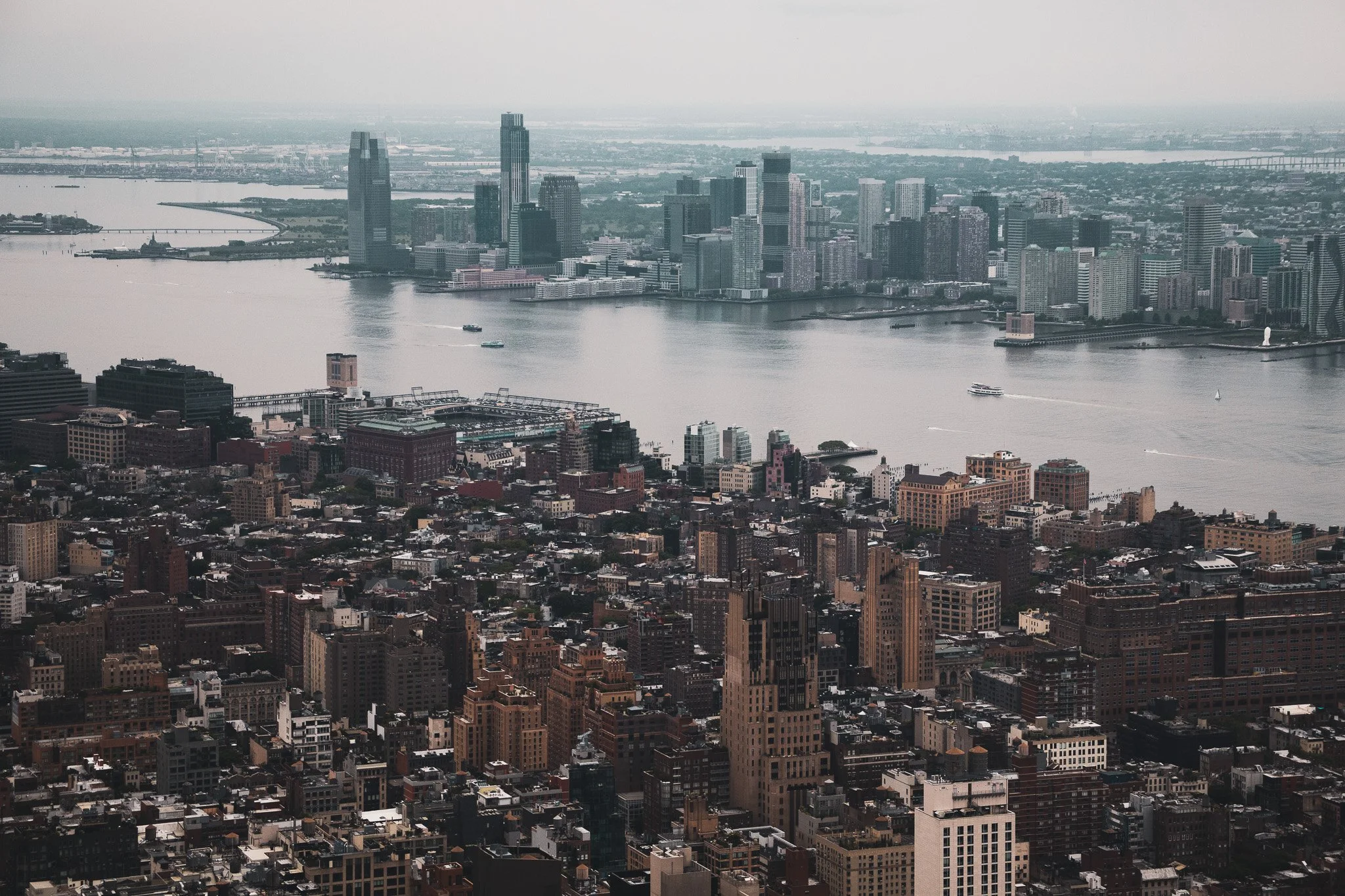 Aerial view of a city skyline with tall skyscrapers and a river with boats, under a cloudy sky.