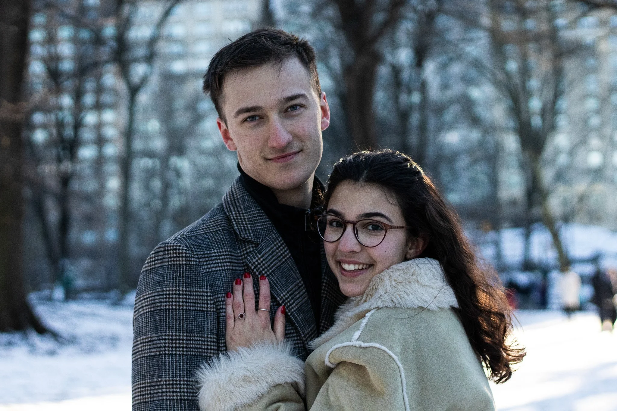 A young man and woman are close together outdoors in a winter setting with bare trees and snow on the ground, smiling at the camera.