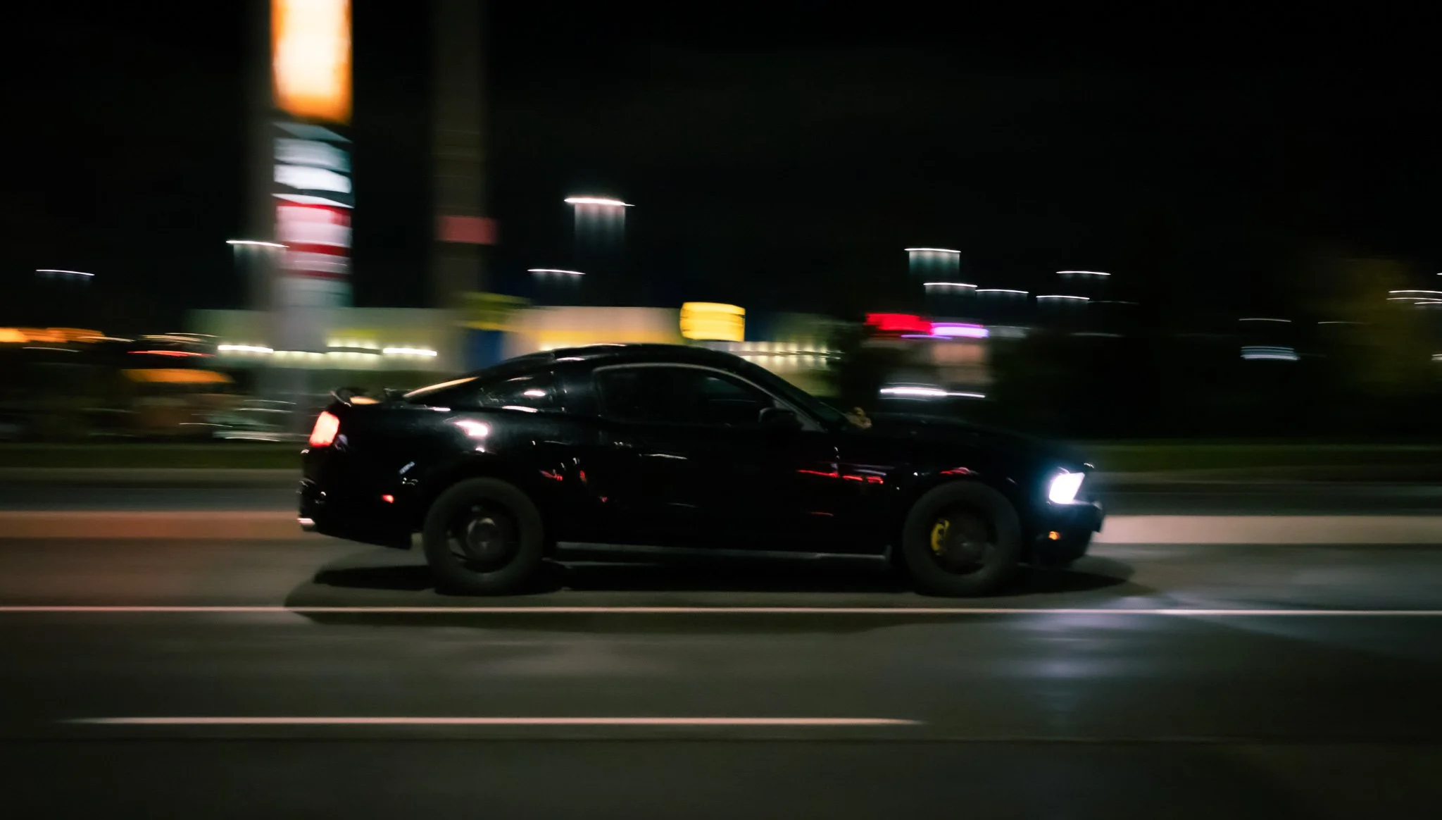 Nighttime speed shot of a black sports car with yellow hubcaps driving on a highway, blurred city lights in the background.