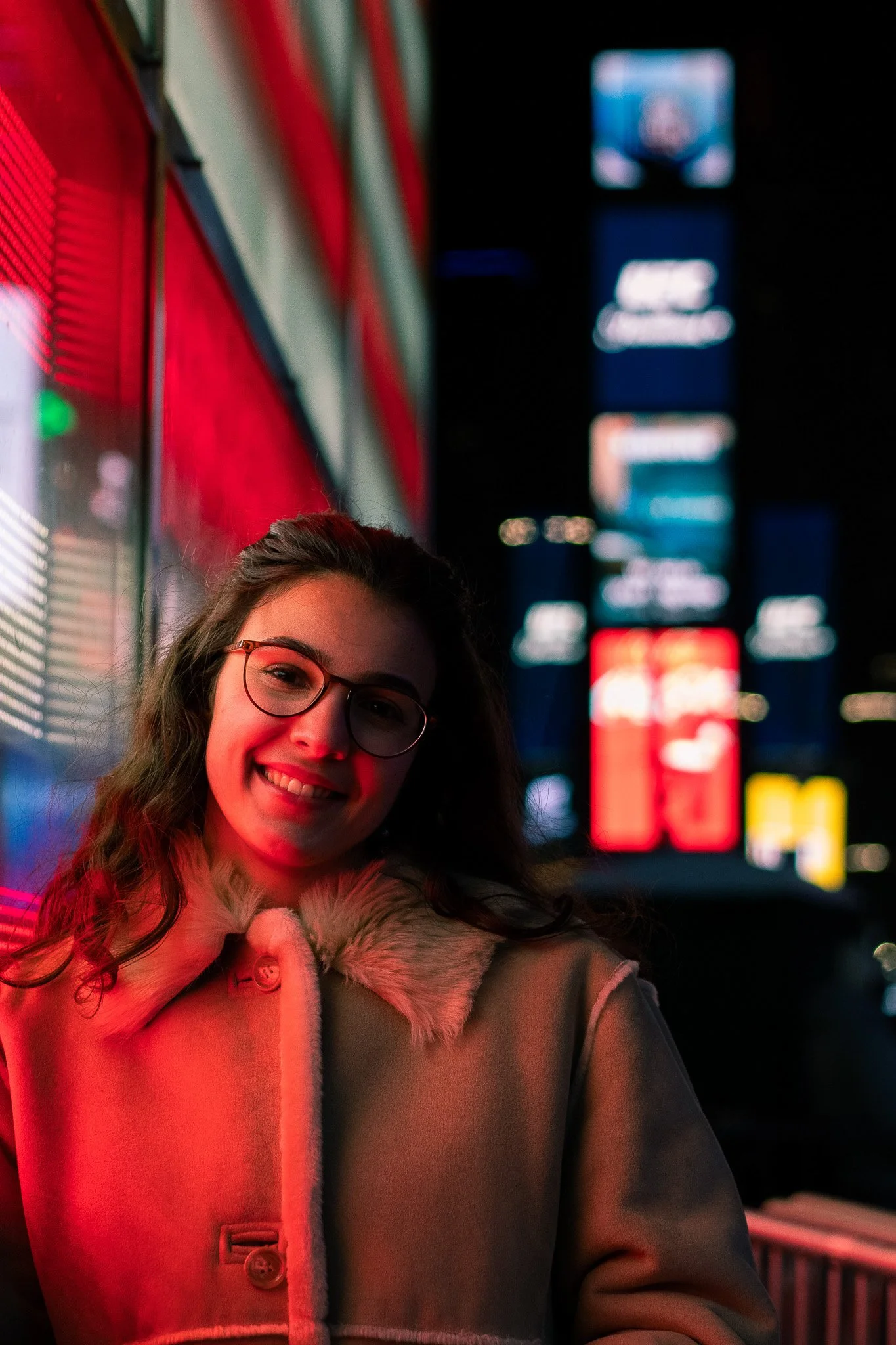 A smiling young woman with glasses and long wavy hair wearing a coat with a fur collar standing in front of brightly lit city billboards at night.