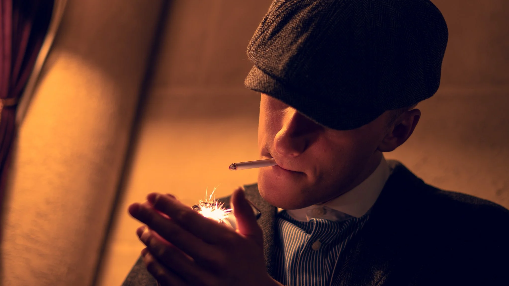 A man wearing a gray hat and a striped shirt lighting a cigarette with a match or lighter, with sparks visible, in a dimly lit setting.
