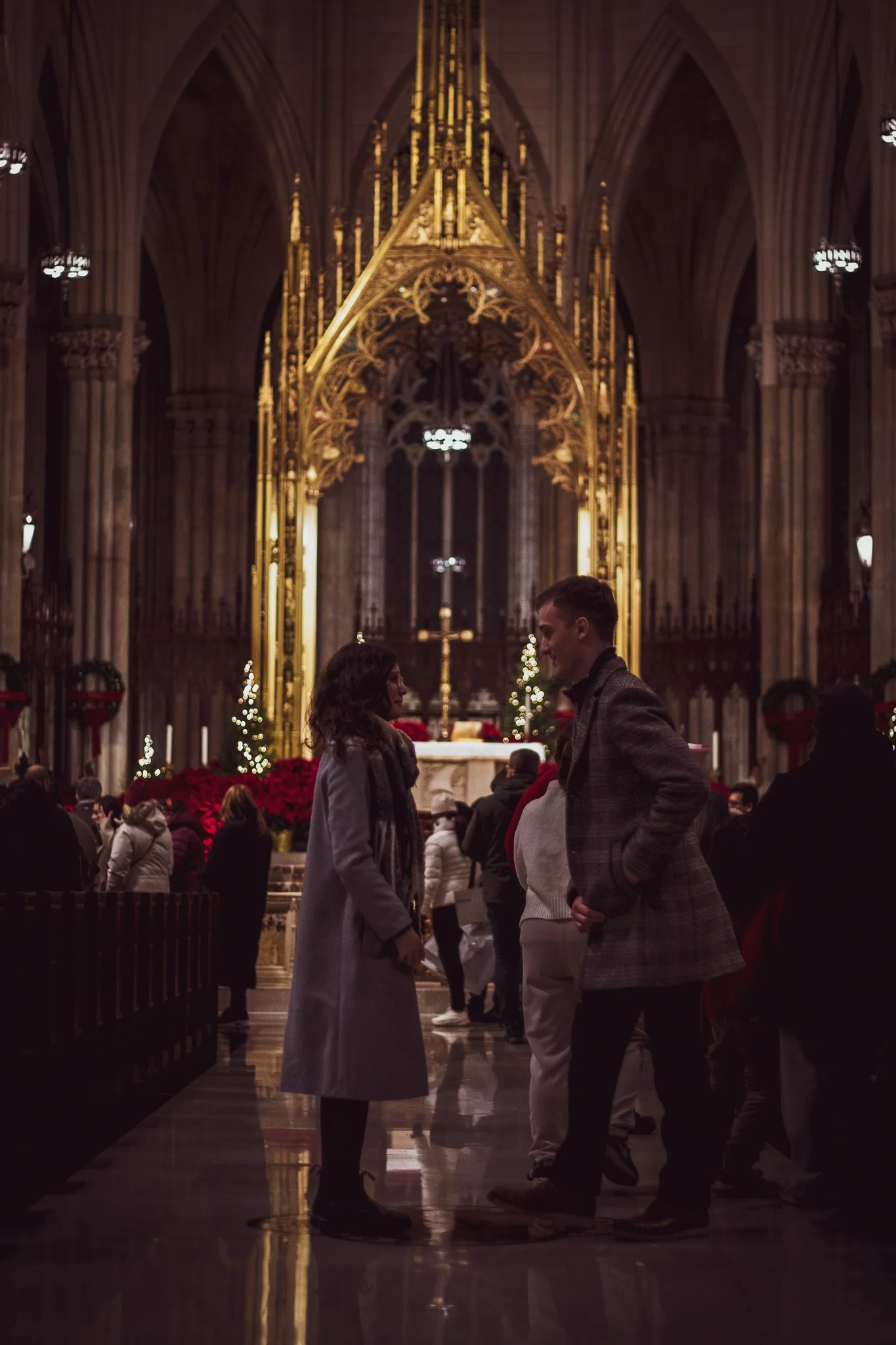 Inside a grand church or cathedral decorated for Christmas, two people stand facing each other, engaged in conversation. The background features tall gothic arches, ornate gold decorations, Christmas trees, and numerous people seated or standing.