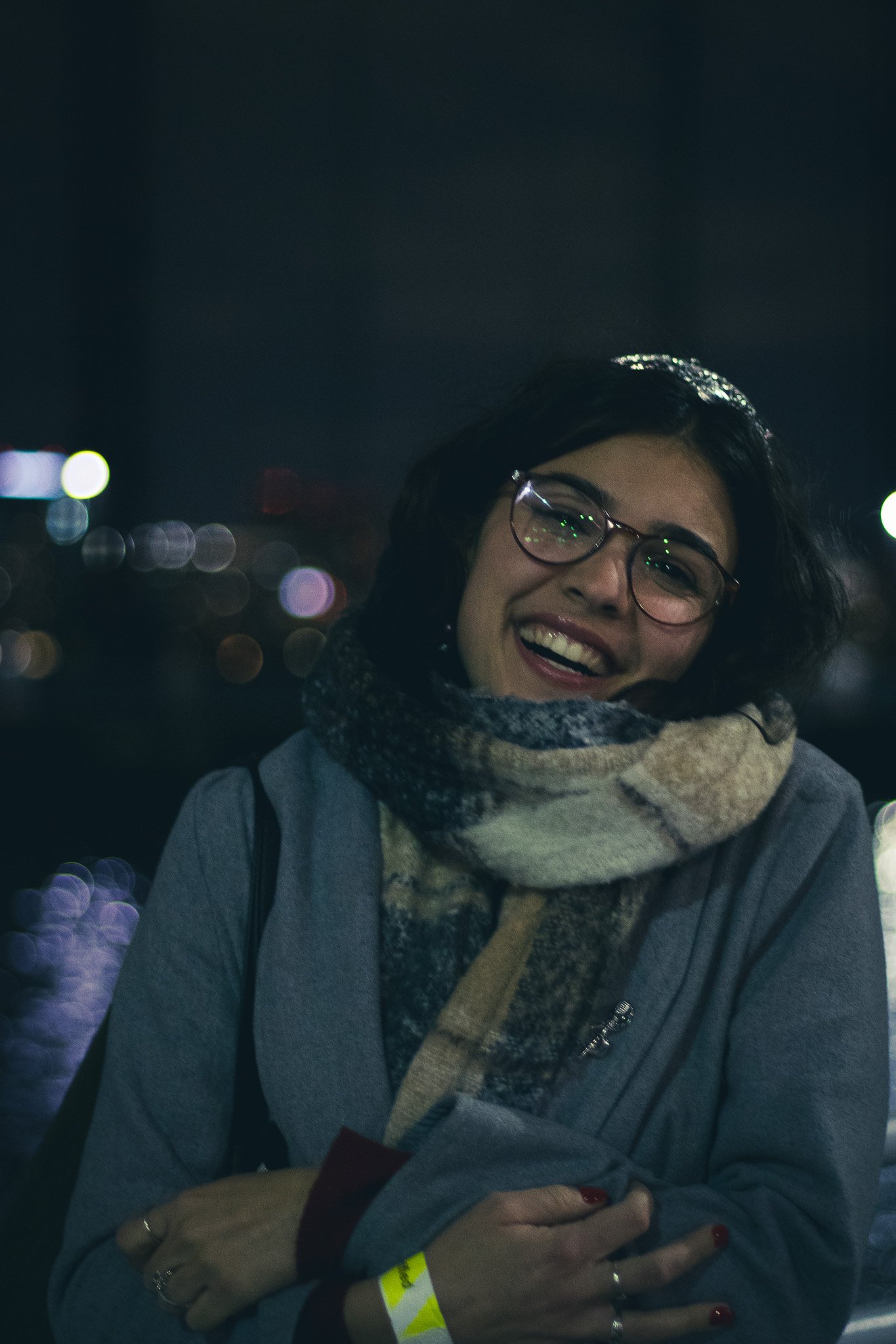 A woman wearing glasses and a scarf, smiling at night with city lights blurred in the background.