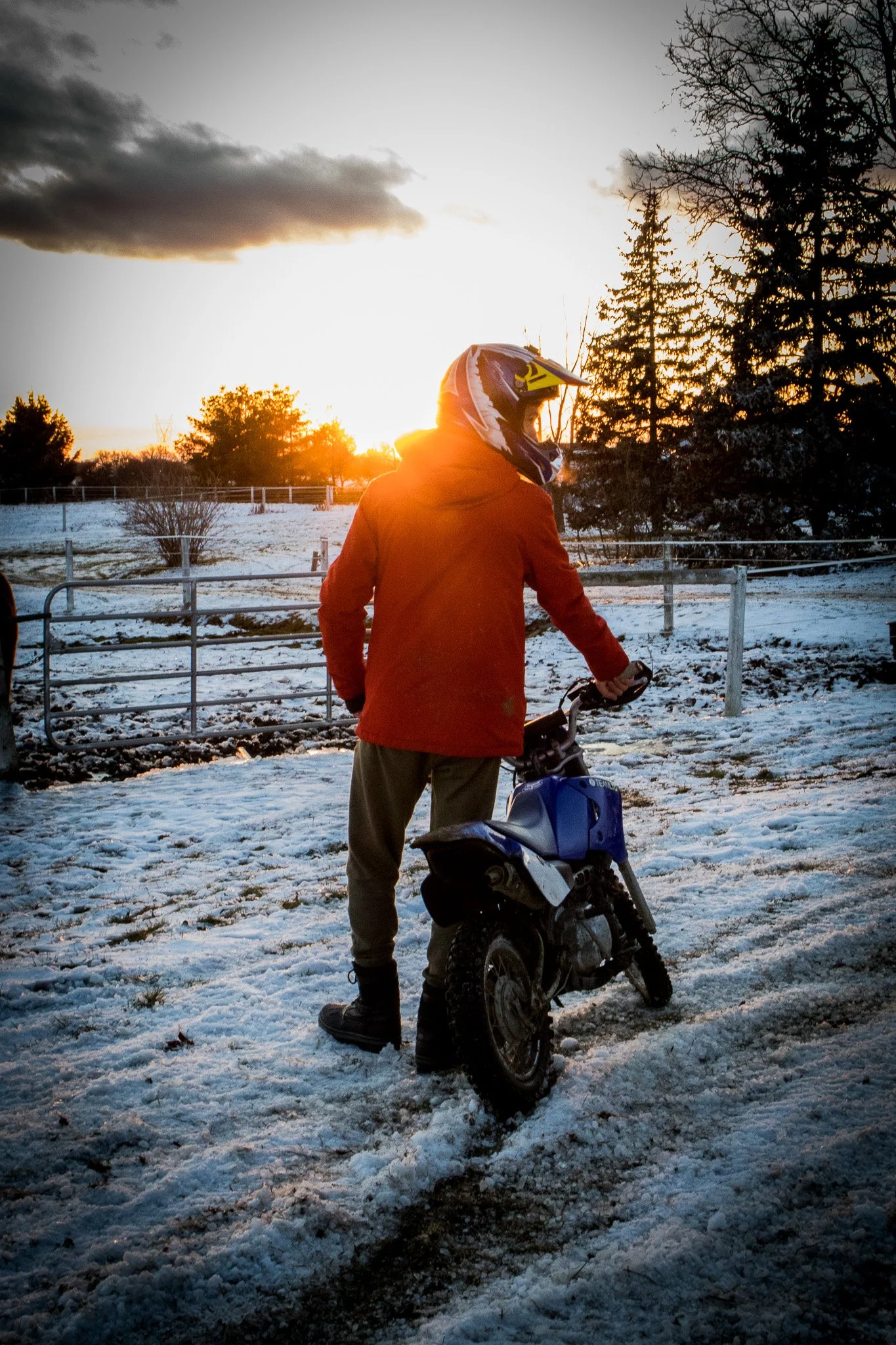 Person wearing an orange jacket and riding a dirt bike on a snowy ground during a sunset, with trees and a fence in the background.