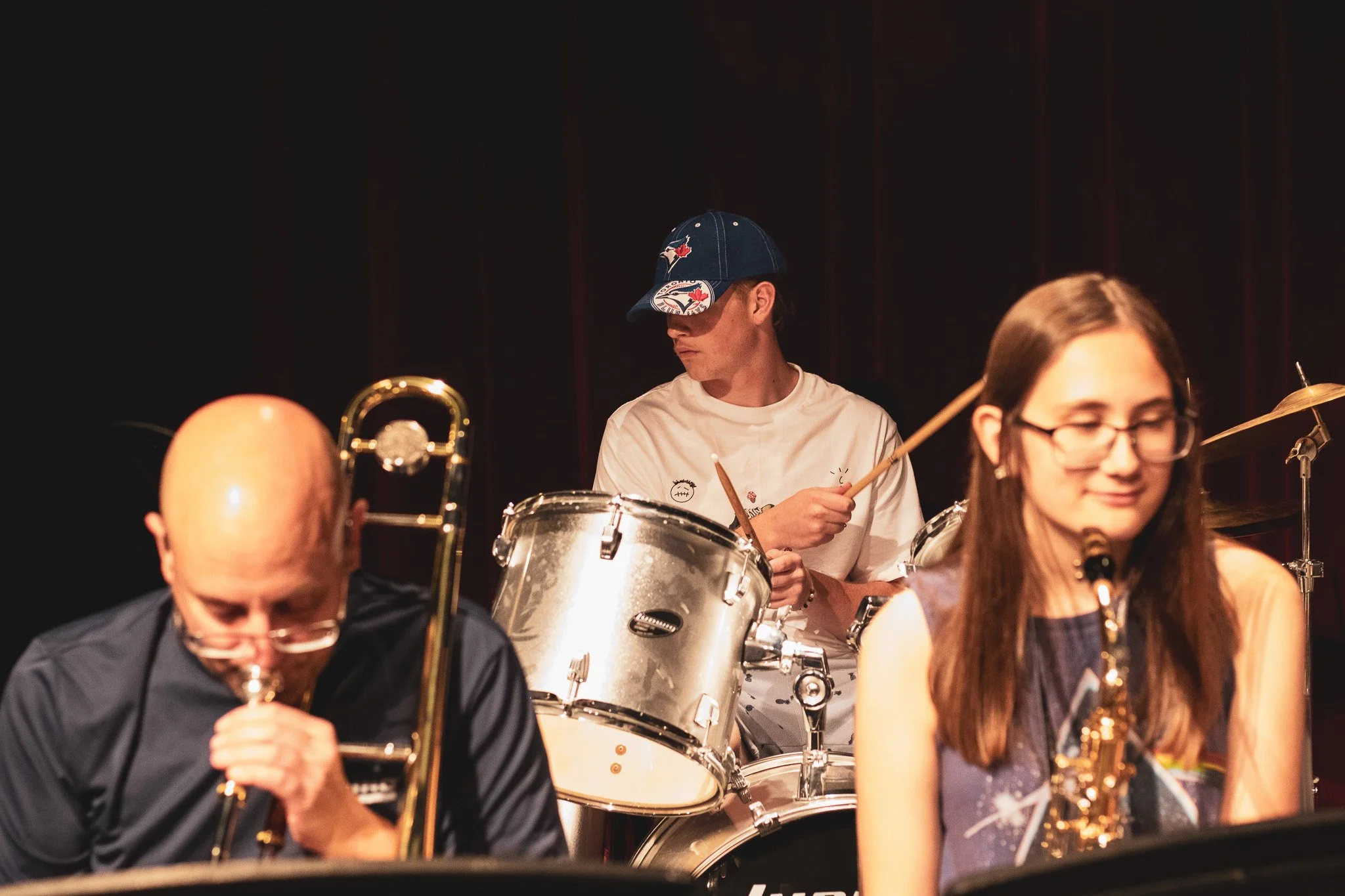 Musicians playing instruments in a band, including a man in a baseball cap playing drums, a bald man with glasses playing a trumpet, and a woman with glasses playing a saxophone.