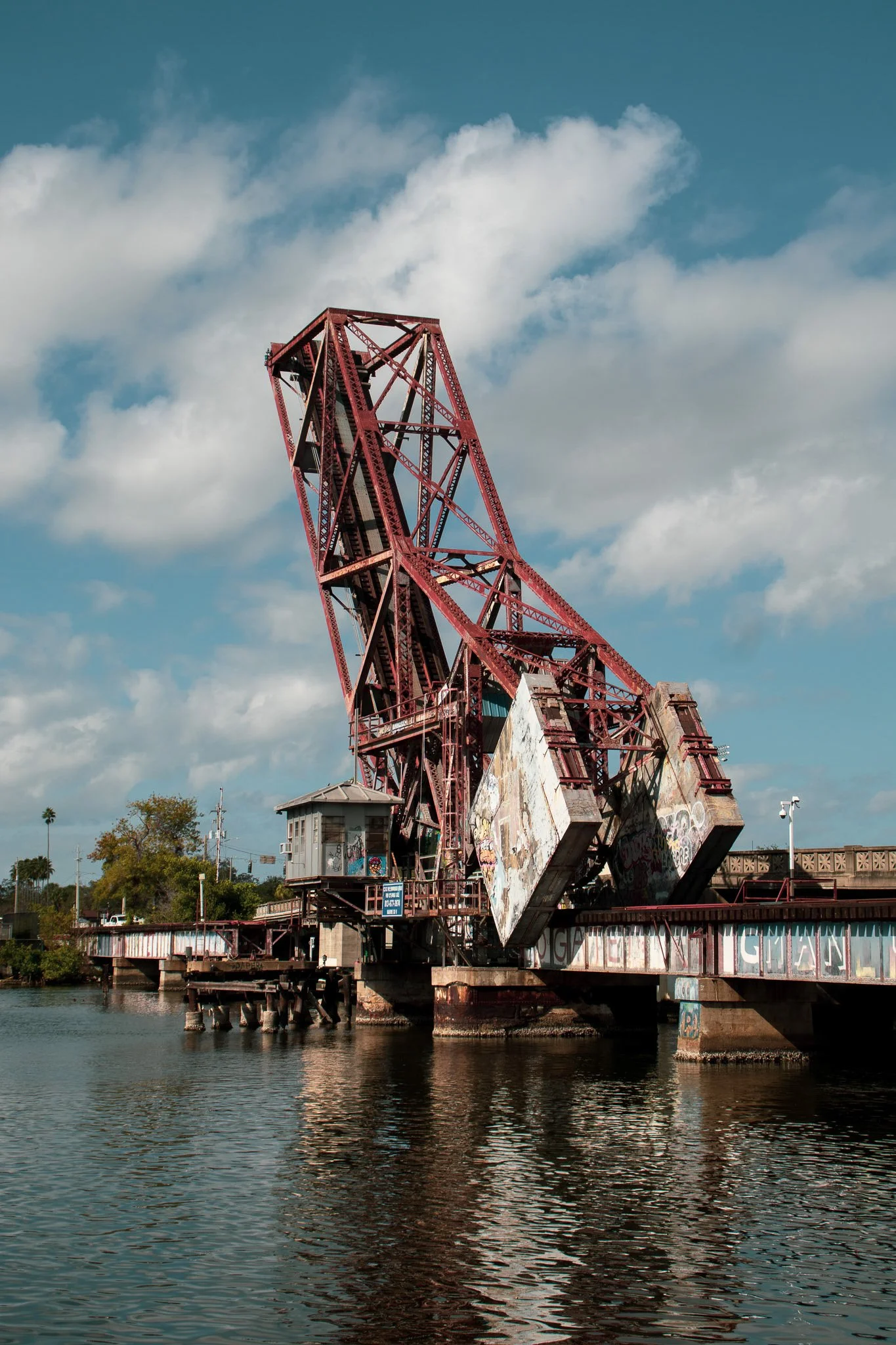 A red, rusted old amusement park dark ride structure on a pier extending over water, with a cloudy blue sky in the background.