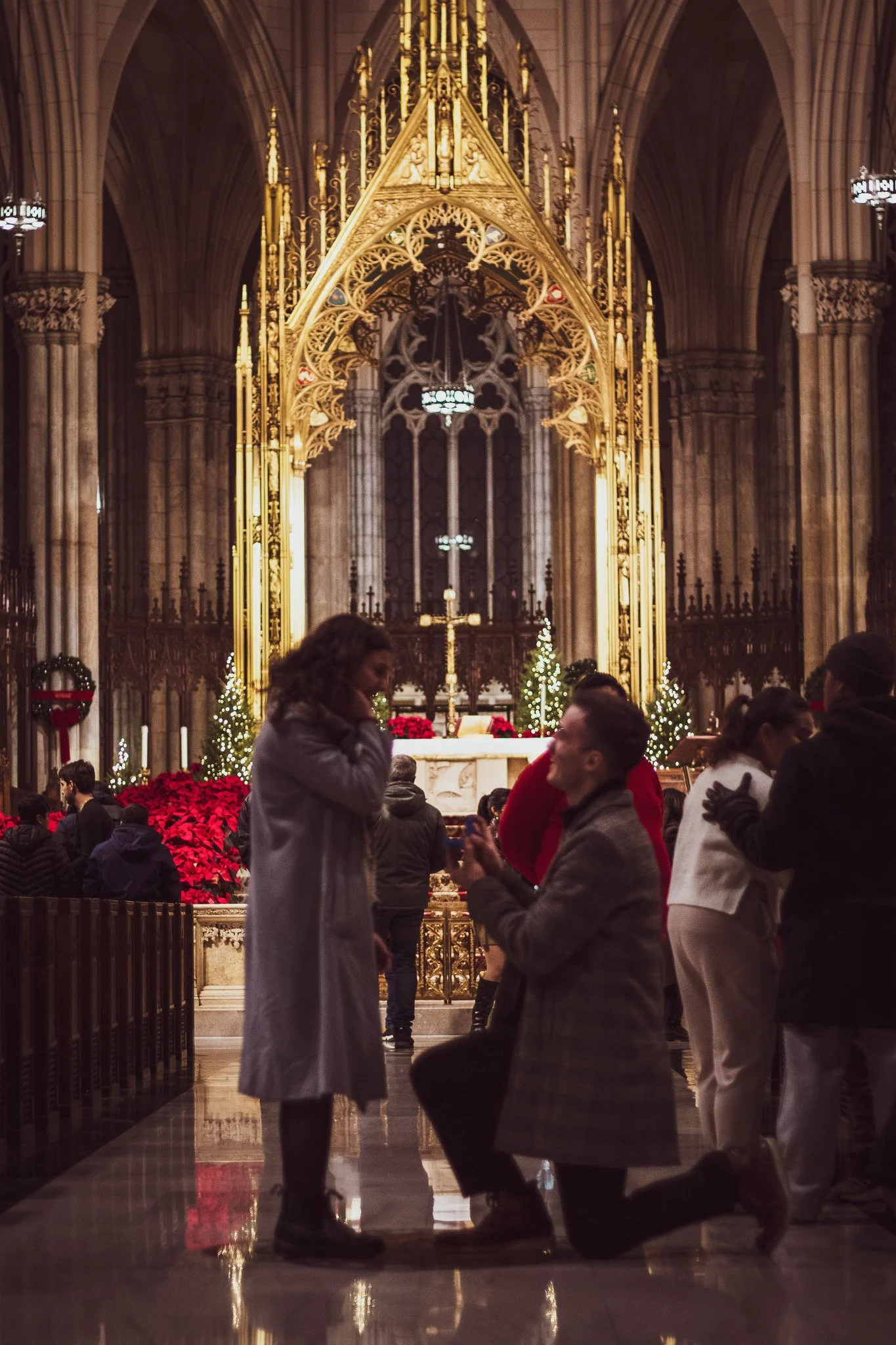 A bishop is kneeling on one knee, proposing to a woman in a church with Gothic architecture decorated for Christmas. The woman is standing and looking down at the man. People are gathered around, some watching the proposal. There are Christmas trees 
