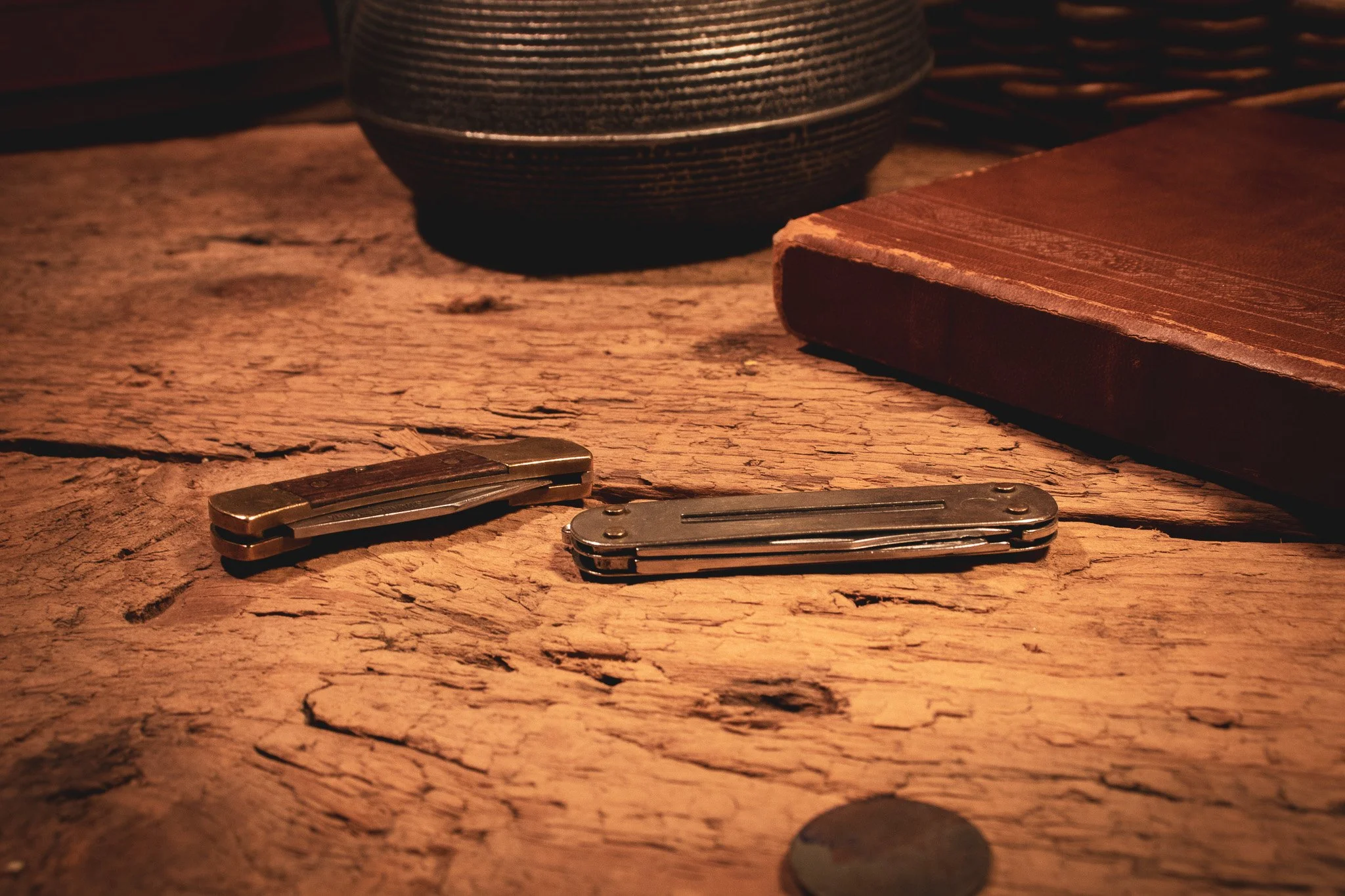 A wooden table with a small black bowl, three pocket knives, a closed book with a red cover, and two coins.